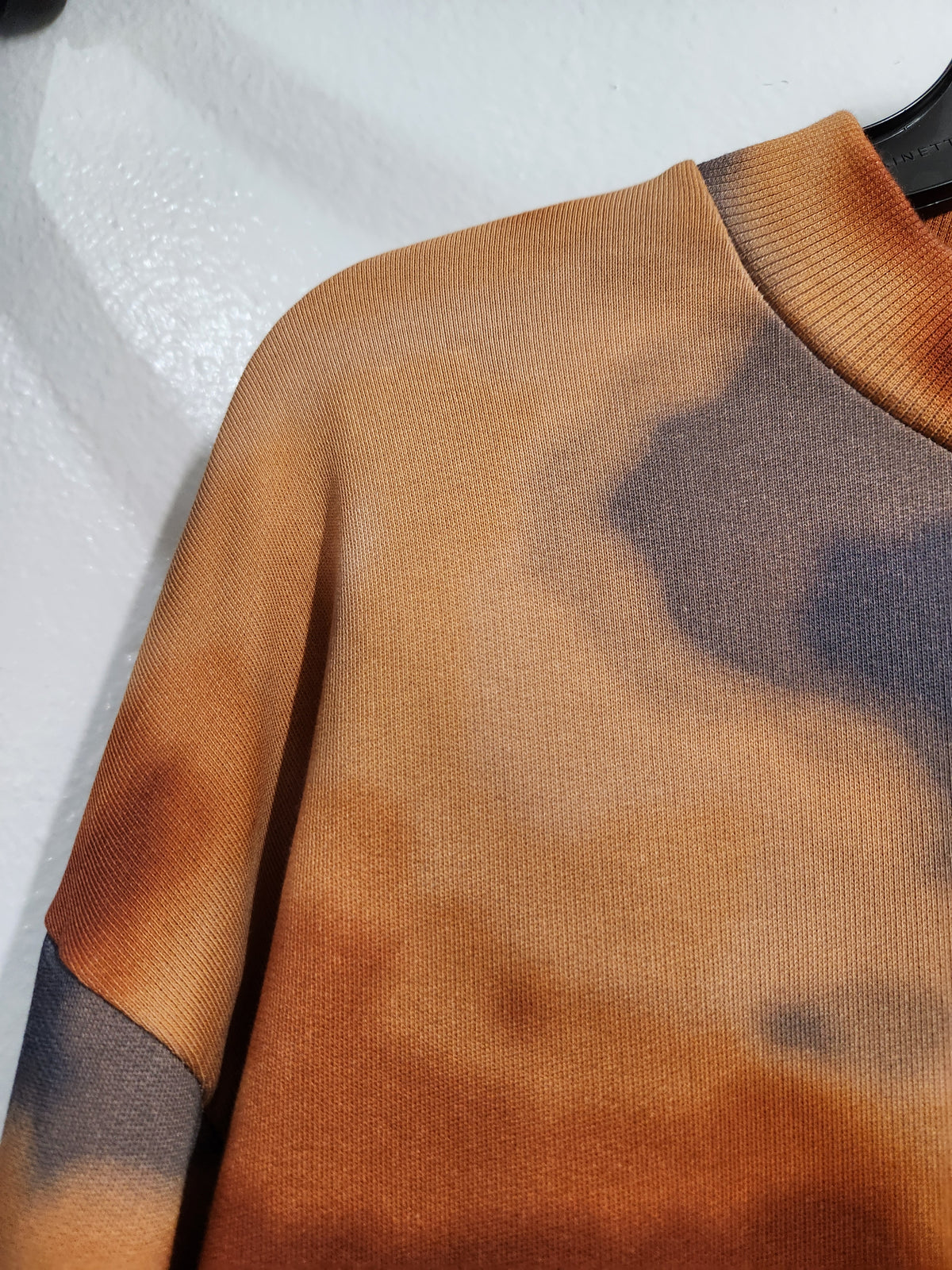 Close-up of a knit crewneck sweater with a mottled tie-dye pattern in warm brown, tan, and dark charcoal gray, shown on a hanger against a light textured wall.