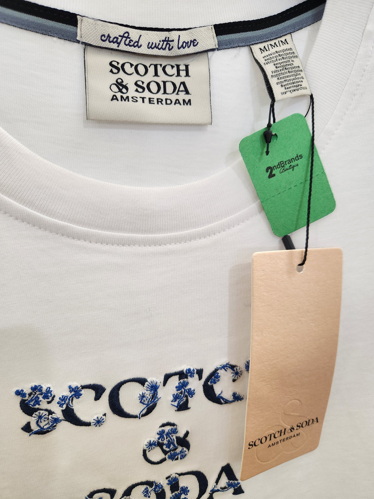 Close-up of a white SCOTCH & SODA t-shirt showing the embroidered brand name in navy blue with blue floral accents, along with neck labels and hang tags.