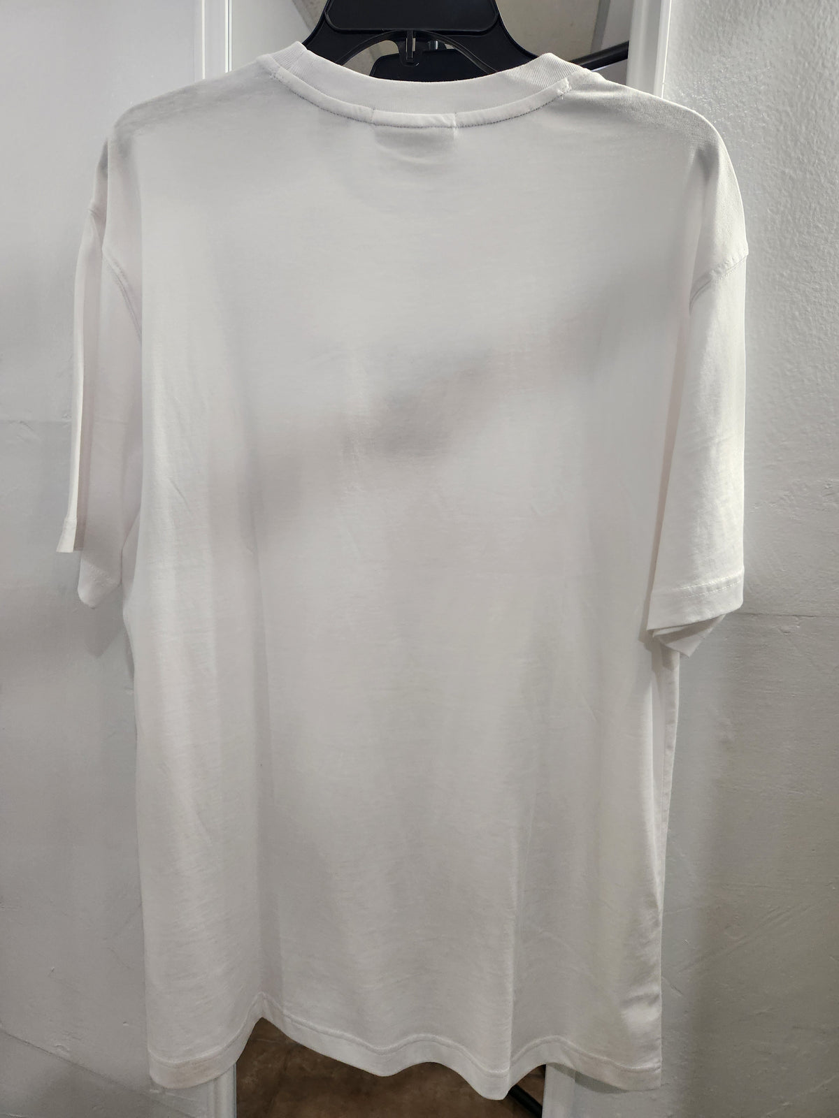 A plain white short-sleeve crewneck t-shirt hanging on a black hanger against a white wall.