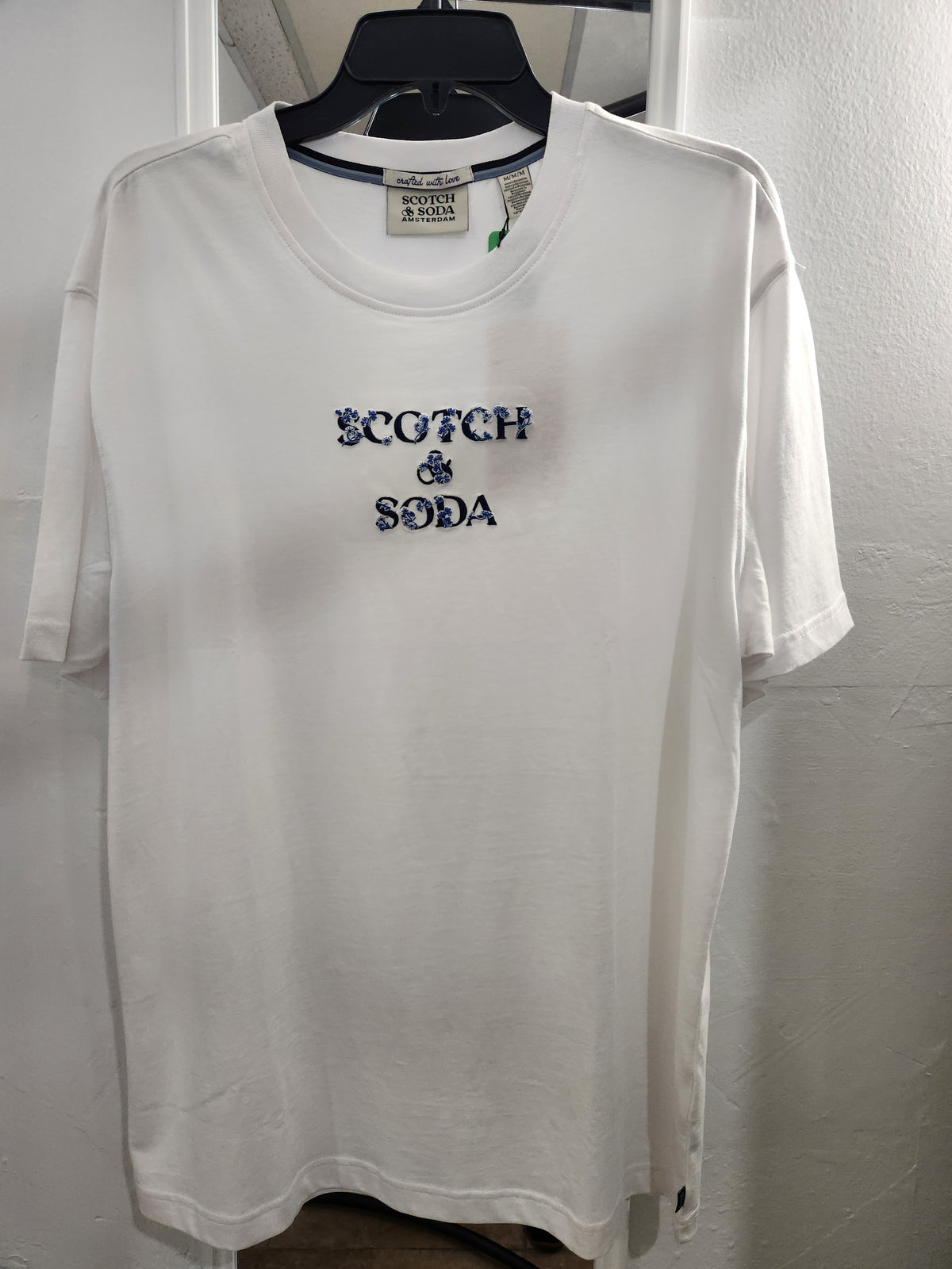 A white short-sleeve crewneck t-shirt hanging on a black hanger against a white wall. The shirt features the text 'SCOTCH & SODA' printed in navy blue on the center chest.