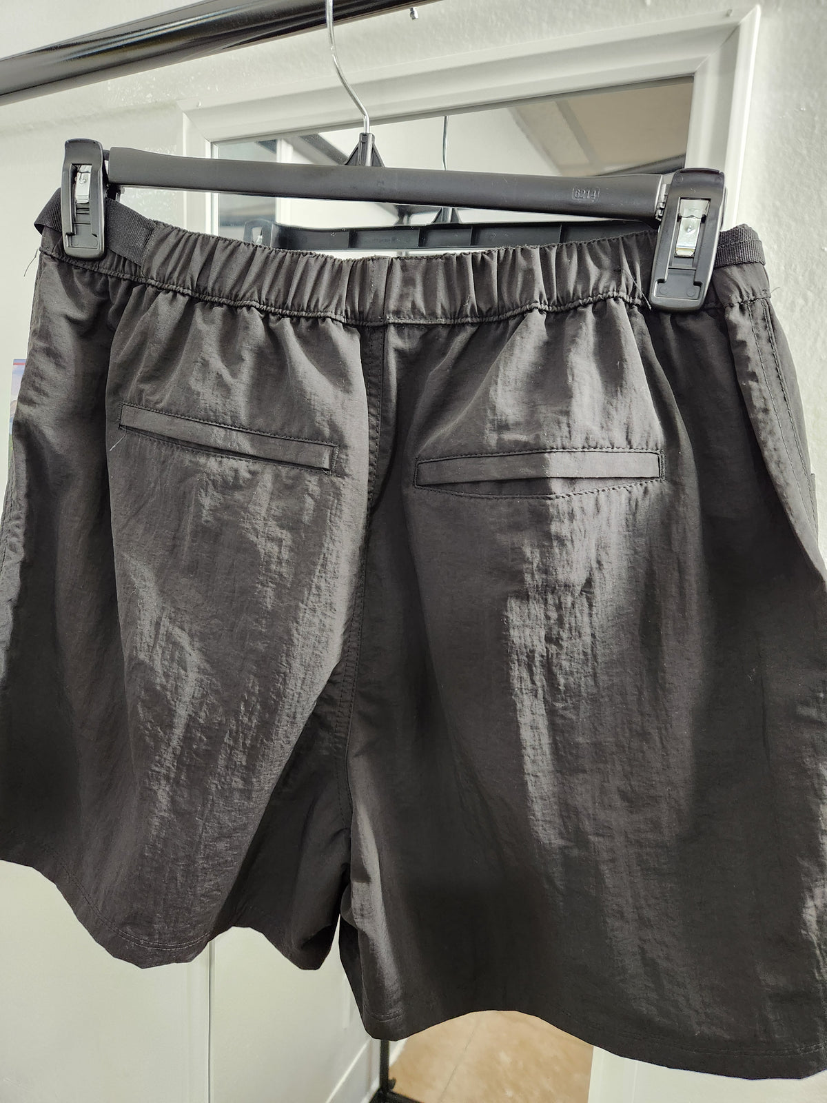 Dark gray elastic-waist shorts with two front patch pockets, hanging on a black hanger against a white door frame.