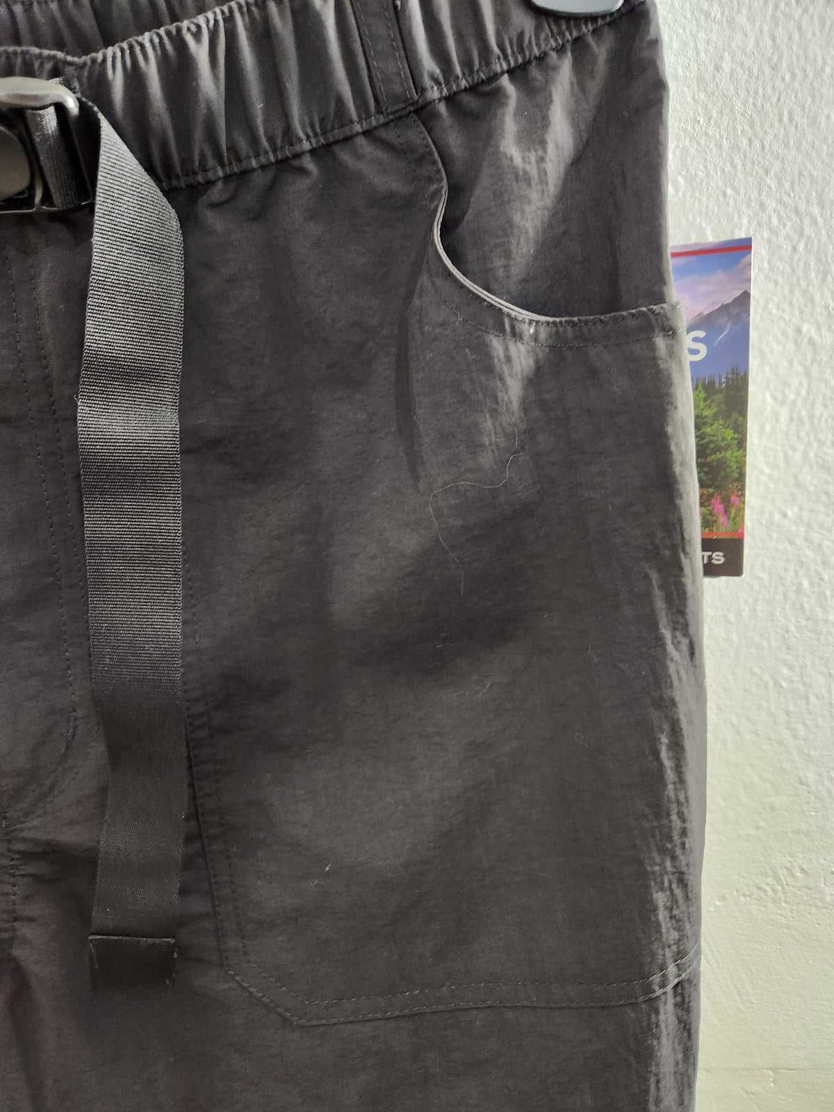 Close-up of dark gray outdoor pants showing the elastic waistband, a vertical drawstring, and a side pocket against a white wall.