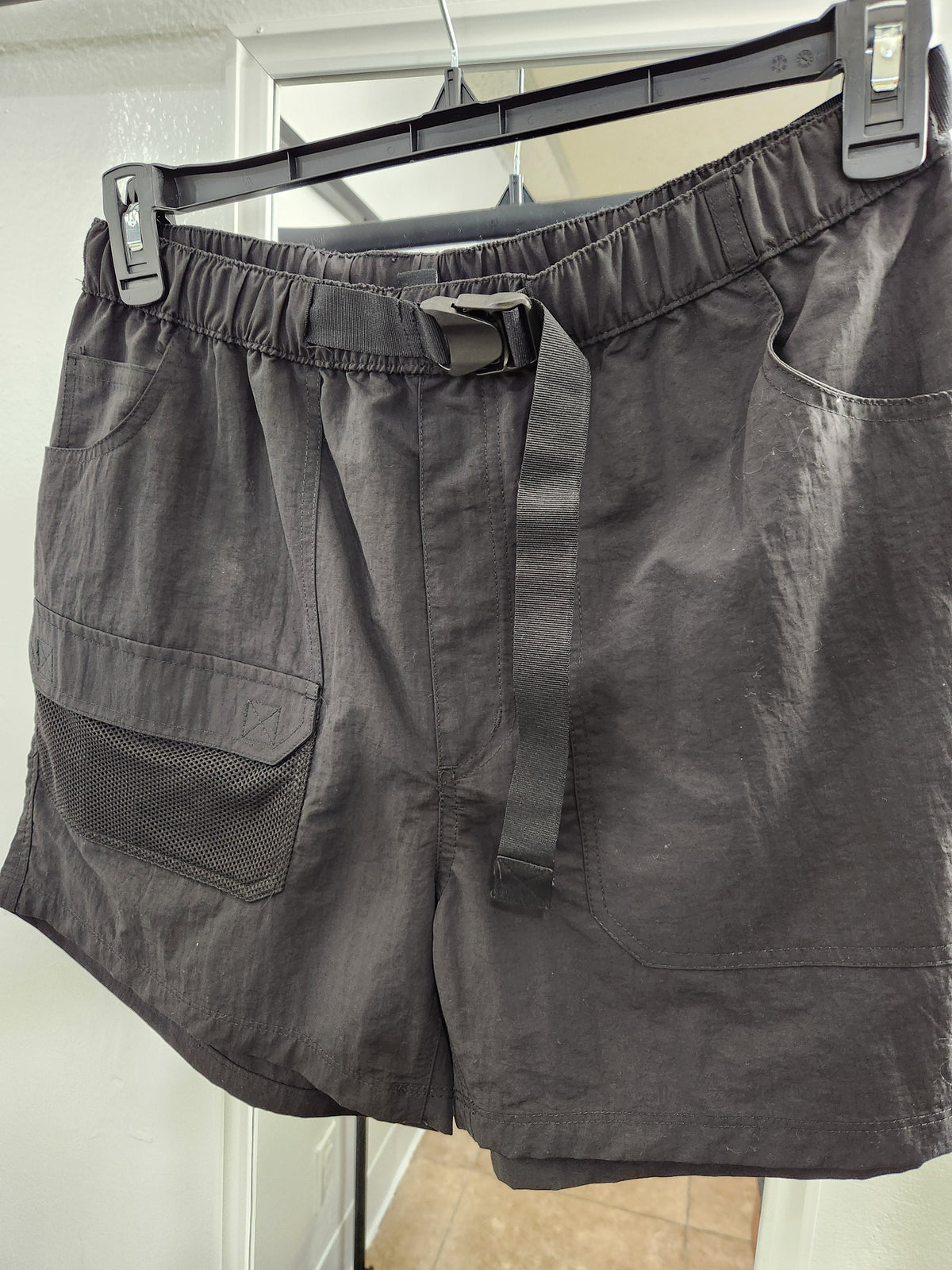 Dark gray cargo shorts with an elastic waistband and a matching fabric drawstring, hanging on a black hanger against a white door background.