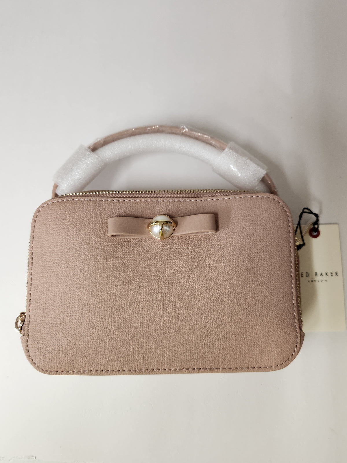 A small structured handbag in light blush pink pebbled leather with a top handle wrapped in protective white foam, a matching bow with a pearl-like center, and a hangtag from Ted Baker London, photographed against a plain off-white background.