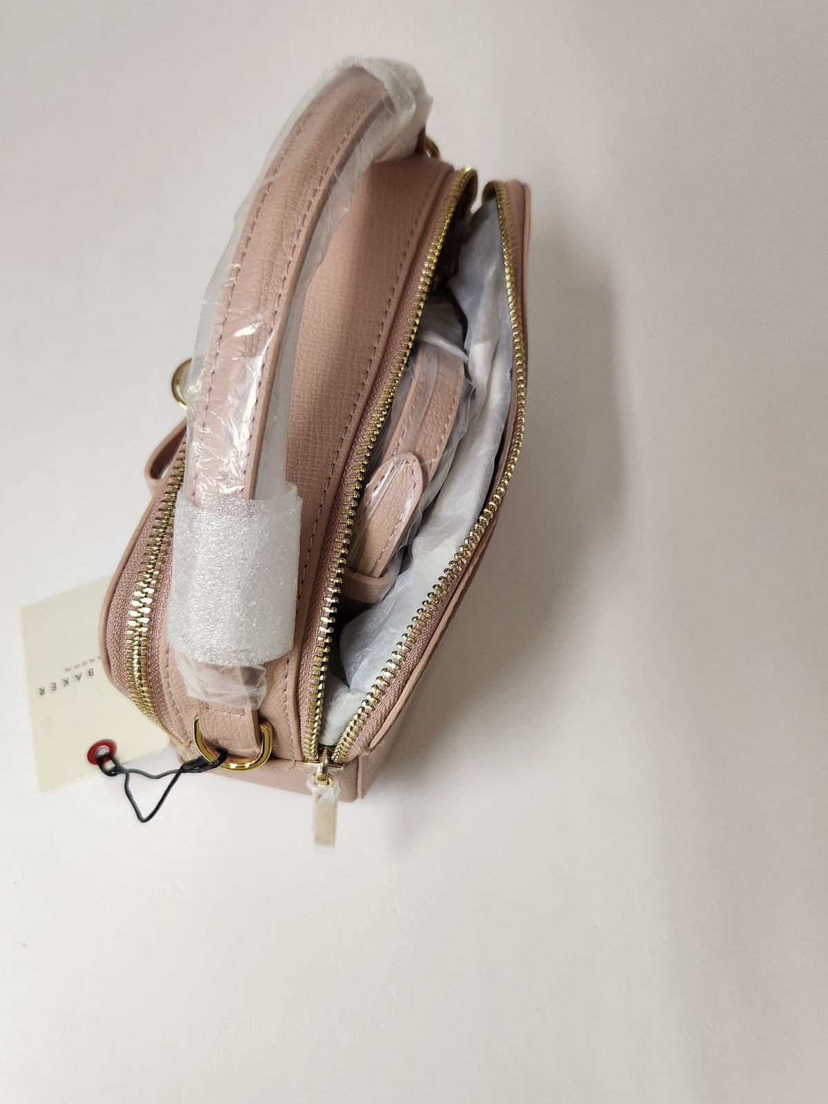 A blush pink textured leather cosmetic pouch with gold zippers, shown open to reveal its interior compartments. The handle is still wrapped in protective plastic, and a small paper tag is attached on the left, set against a plain off-white background.