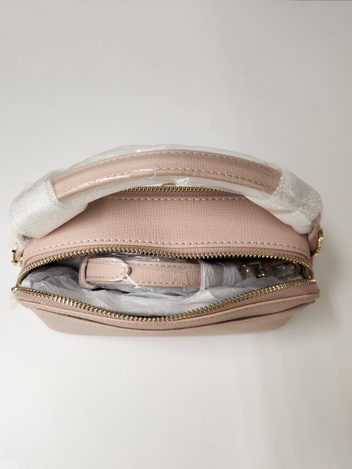 A light pink pebbled leather cosmetic pouch shown open, revealing its interior and a matching pink strap wrapped in protective white paper. The bag features gold-tone zippers and is photographed from a top-down perspective against a clean, off-white background.