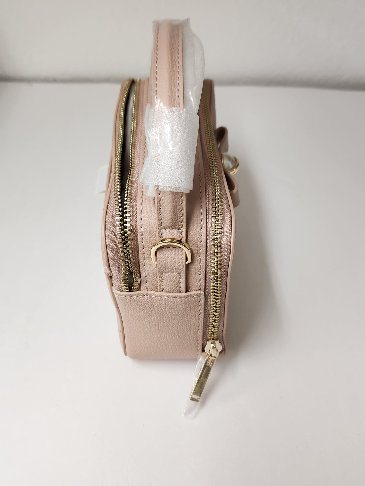 A small blush pink pebbled leather crossbody bag with gold-tone zippers and hardware, shown standing upright on a white surface with protective plastic wrapping still attached to the handle.