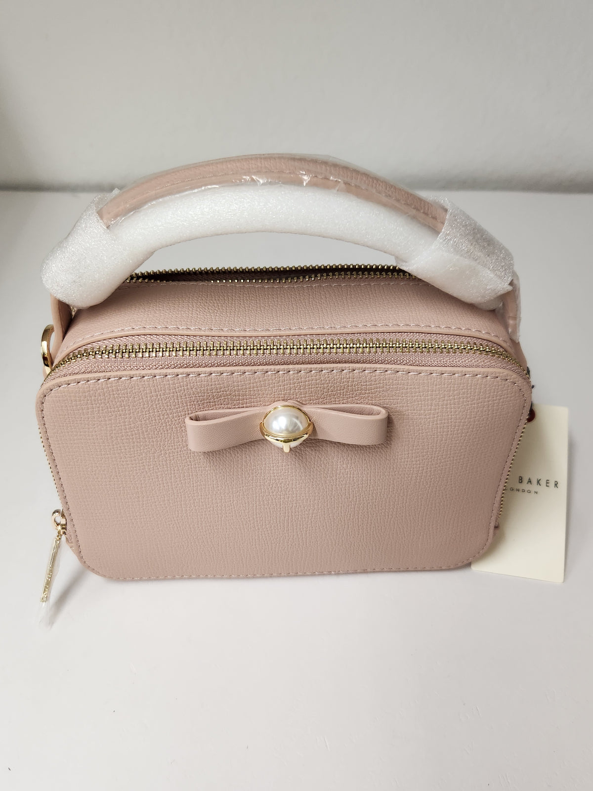 A light pink textured leather cosmetic bag with a top handle wrapped in protective white foam, featuring a small bow with a pearl-like center and gold-tone hardware. A small paper tag with the brand name 'Baker' is visible on the right side.