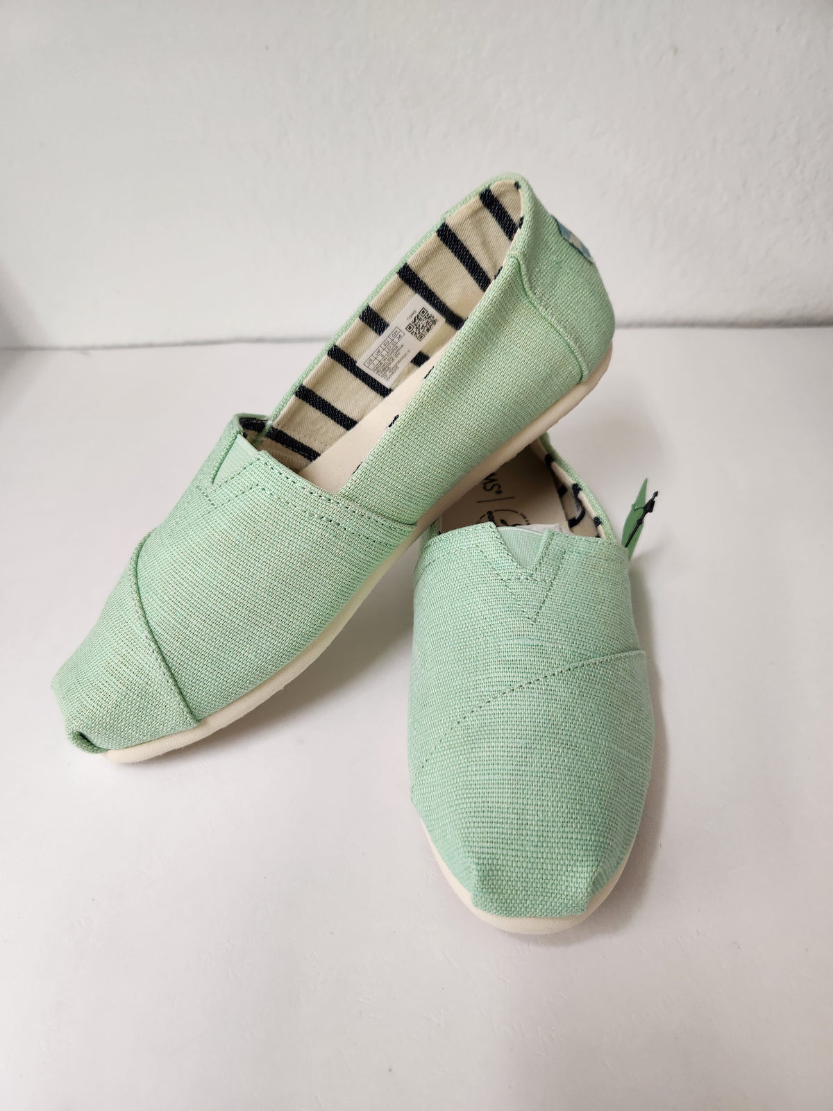 A pair of mint green canvas slip-on shoes with white soles, displayed on a white surface against a white wall.