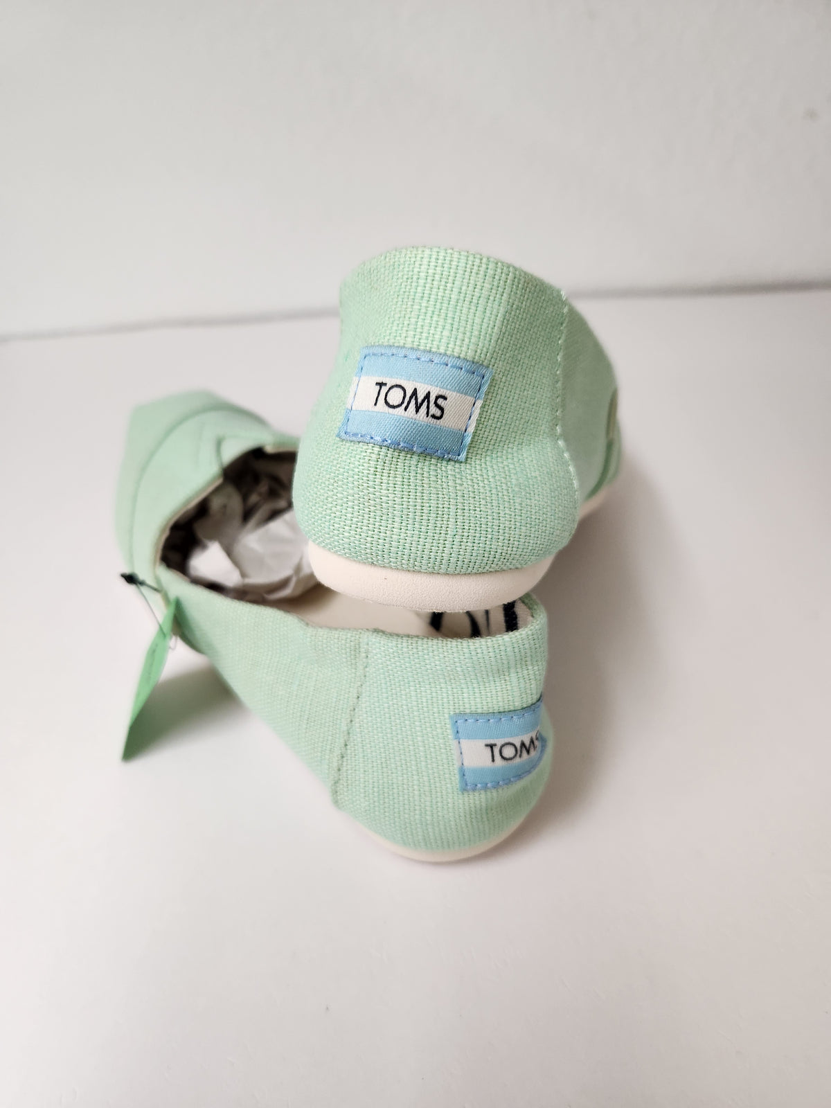 A pair of mint green canvas TOMS slip-on shoes with white soles, displayed on a white surface against a white wall.