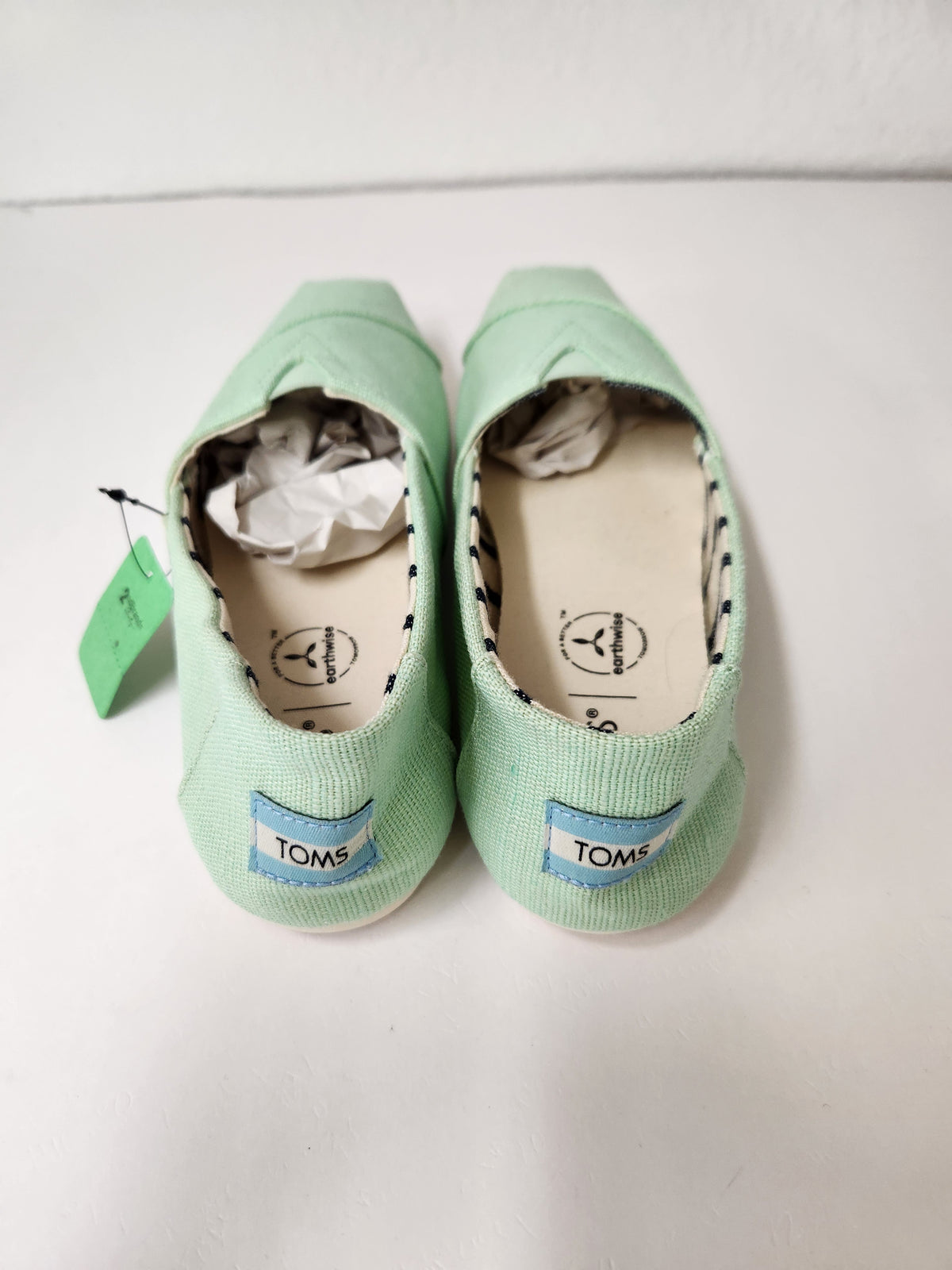 A pair of mint green canvas slip-on shoes by TOMS, shown from the back with their tags and tissue paper still attached, on a white surface.