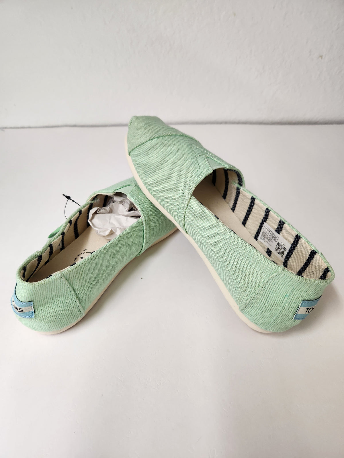 A pair of mint green canvas slip-on shoes, likely from the brand TOMS, displayed on a white surface against a white wall. One shoe is positioned upright to show the striped interior lining, while the other lies on its side showing the textured canvas exterior and the small blue brand tag near the heel.