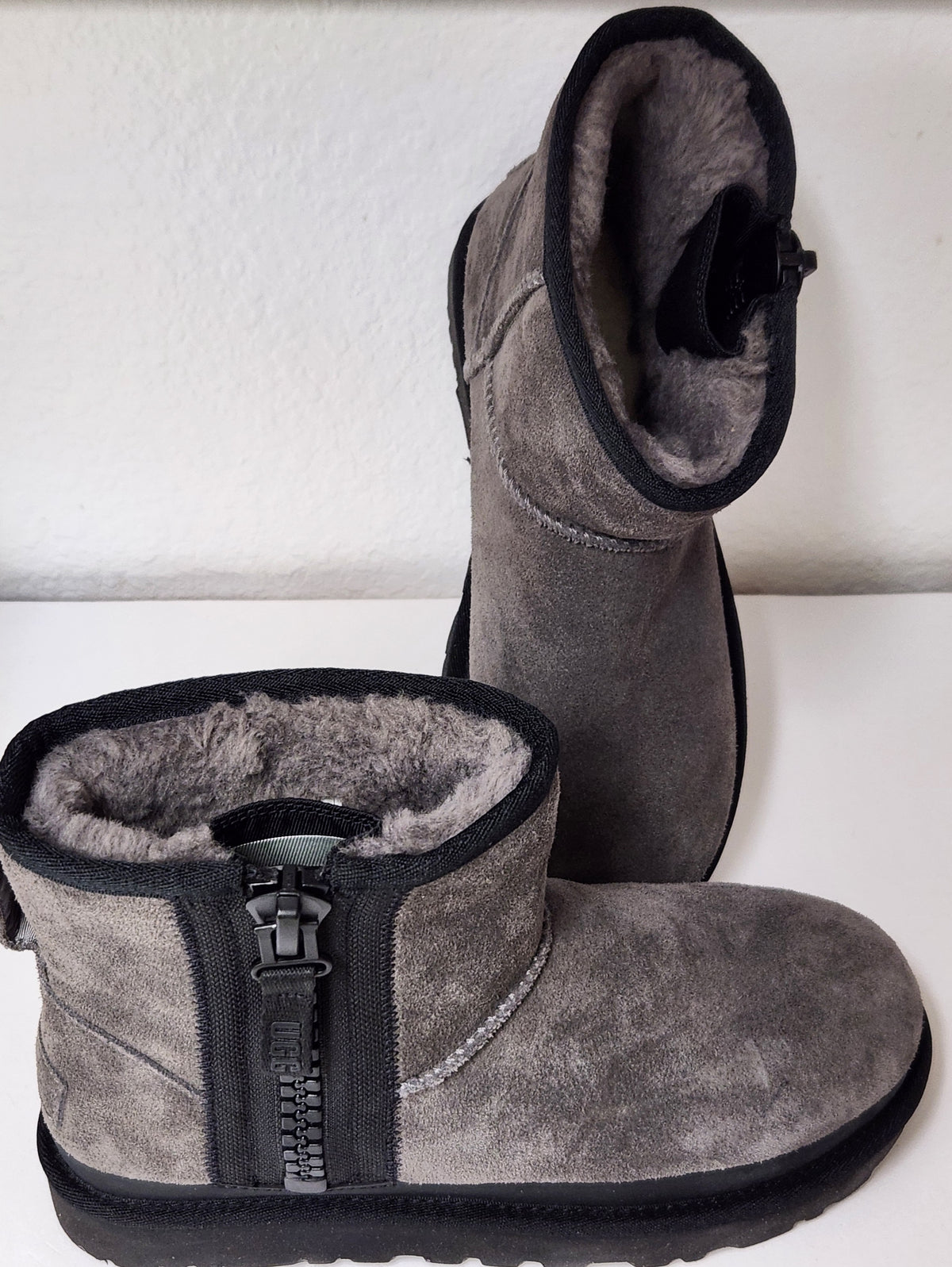 A pair of gray suede ankle boots with black trim and a plush gray shearling lining, featuring a side zipper on the front boot.