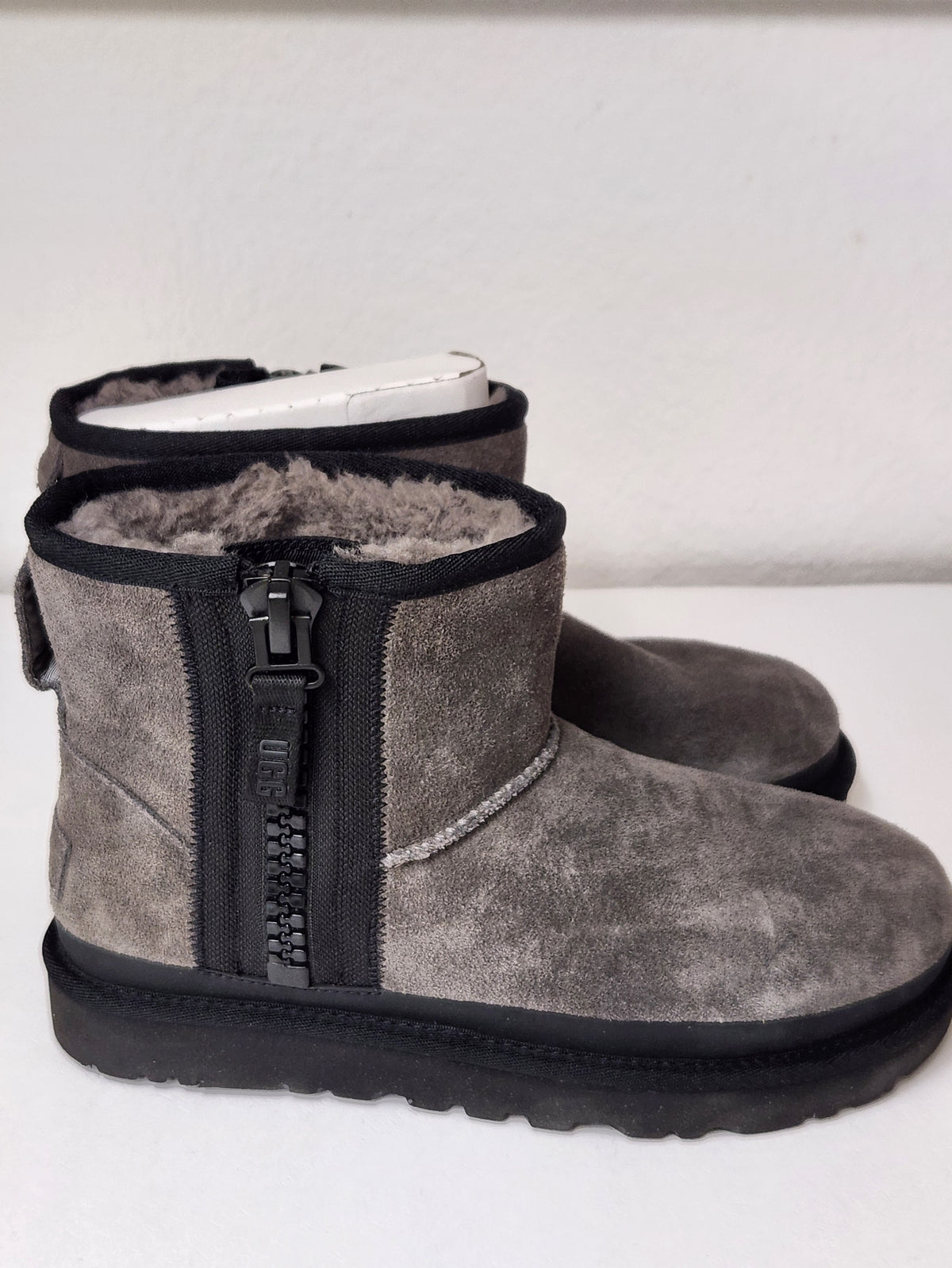 A pair of gray suede ankle boots with black side zippers, black soles, and gray shearling lining, displayed on a white surface against a white wall.