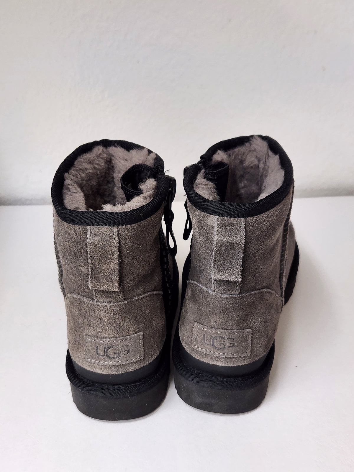 A pair of gray suede UGG boots with black soles and a light gray shearling lining, shown from the back on a white surface against a white wall.