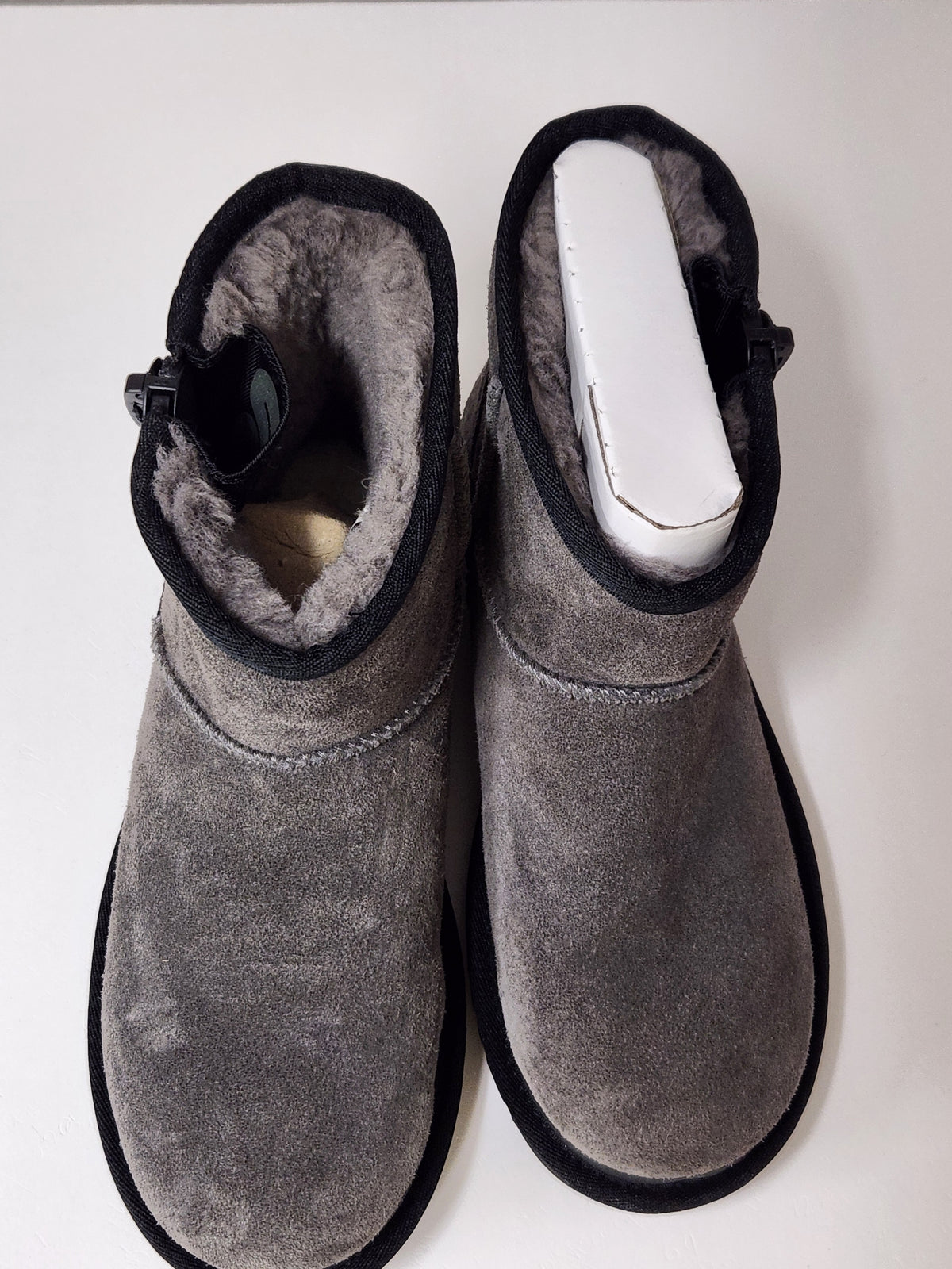 A pair of gray suede UGG boots with black soles and a light gray shearling lining, shown from the back on a white surface against a white wall.