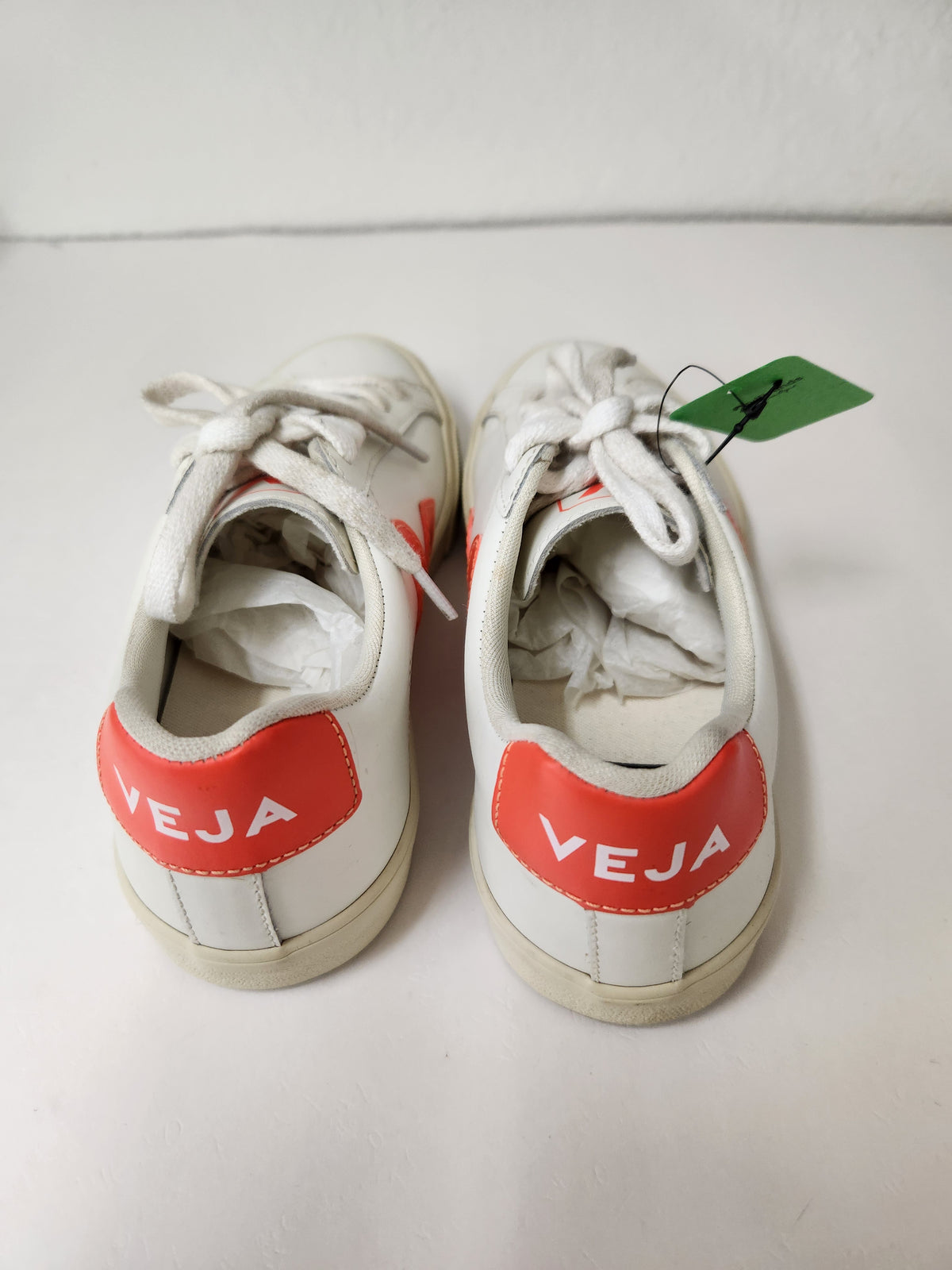 A pair of white leather sneakers with red heel tabs featuring the white text 'VEJA', shown from a top-down view on a white surface with a white background.