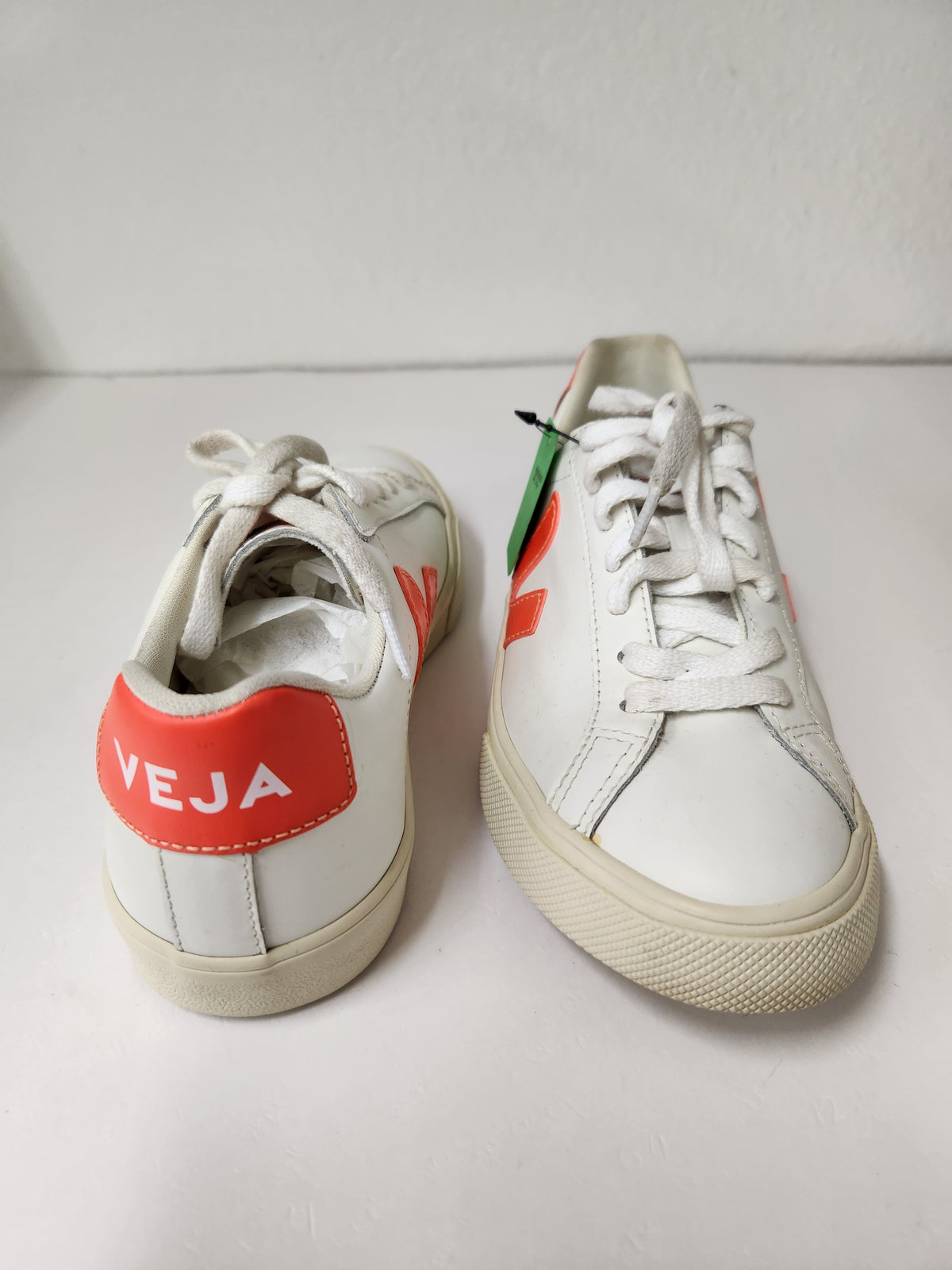 A pair of white leather sneakers with orange-red heel tabs and side accents, featuring the word 'VEJA' in white on the left heel tab. One shoe is shown from the back and the other from the side, on a light-colored surface against a white background.