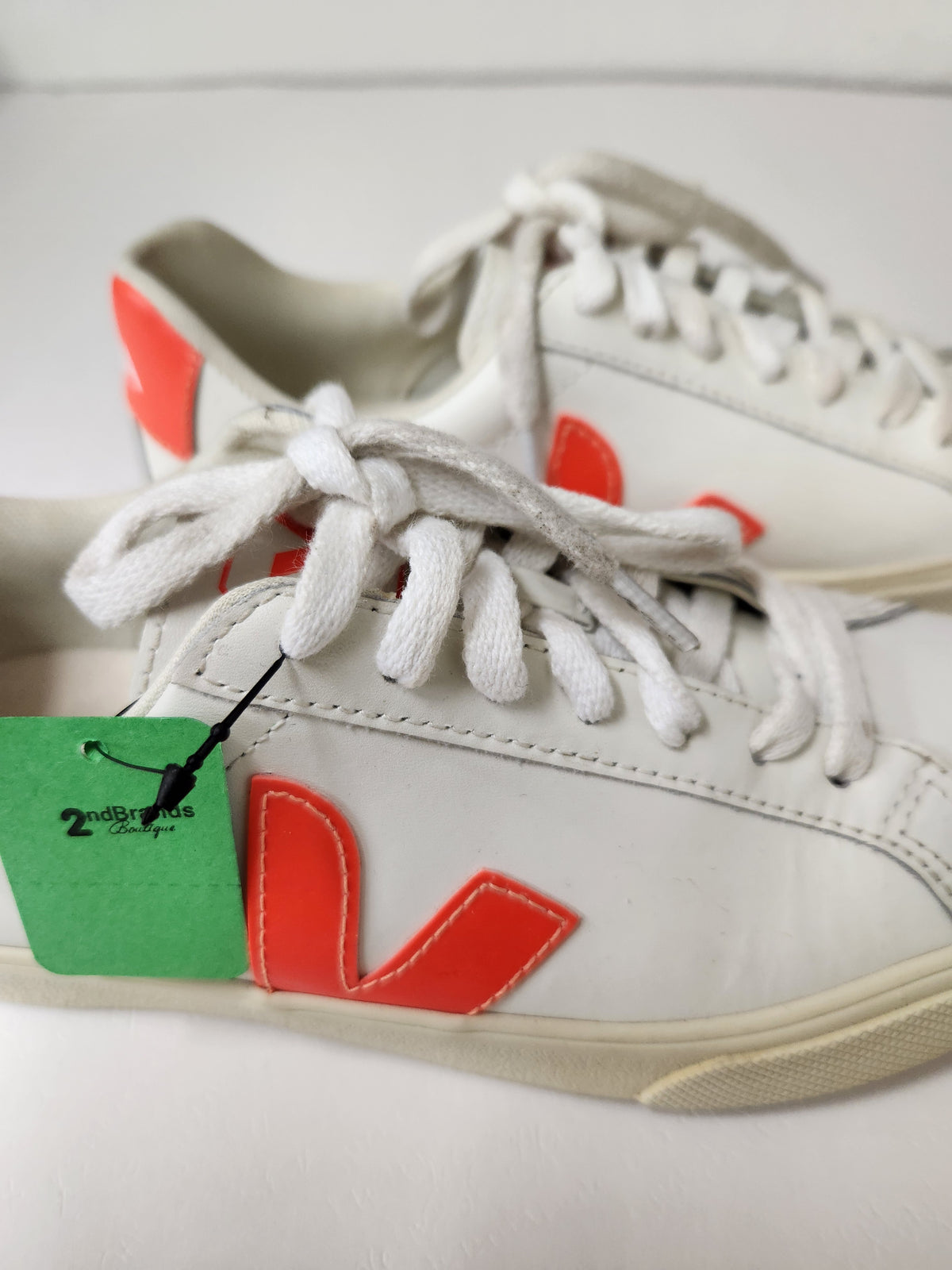 A pair of white leather low-top sneakers with a prominent orange-red 'V' logo on the side, white laces, and a green price tag attached to the shoe in the foreground.