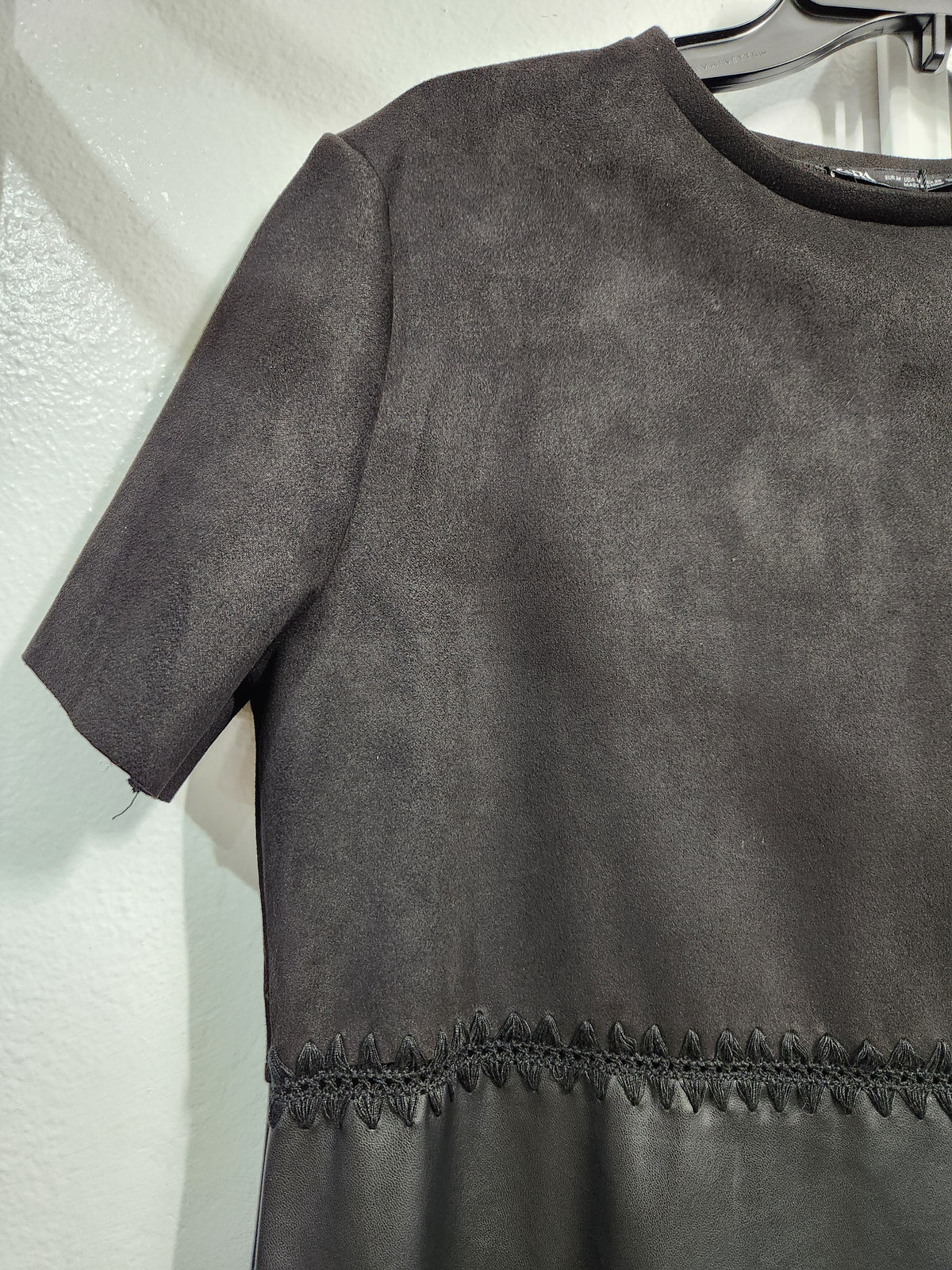 A dark gray short-sleeve top or dress made of textured faux suede, hanging on a black hanger against a white textured wall. The bottom hem features a decorative stitched border.