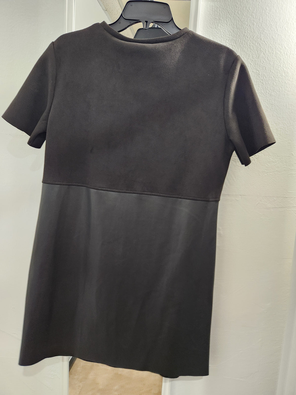 A short-sleeve black dress with a color-blocked design, featuring a textured knit upper half and a smooth faux-leather lower half, hanging on a black hanger against a white wall.