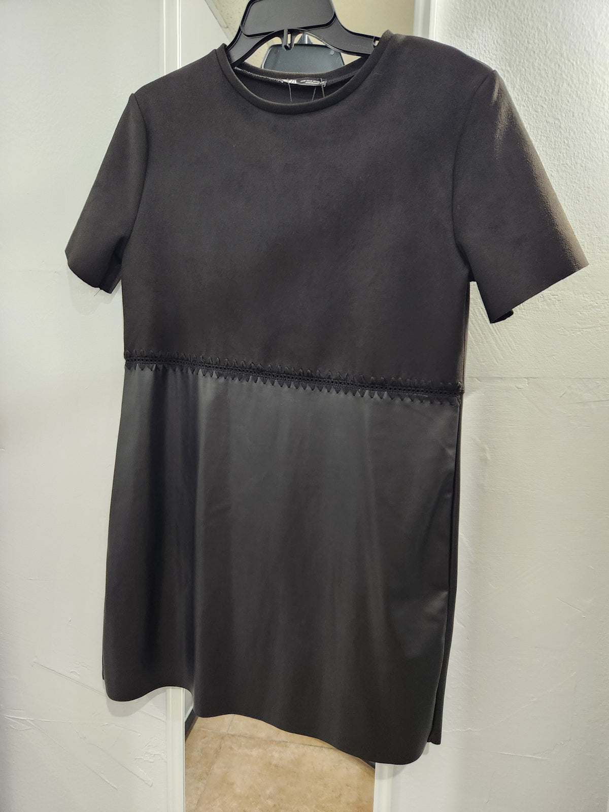 A black short-sleeve dress with a textured, suede-like upper bodice and a smooth faux-leather lower skirt, hanging on a black hanger against a white wall.