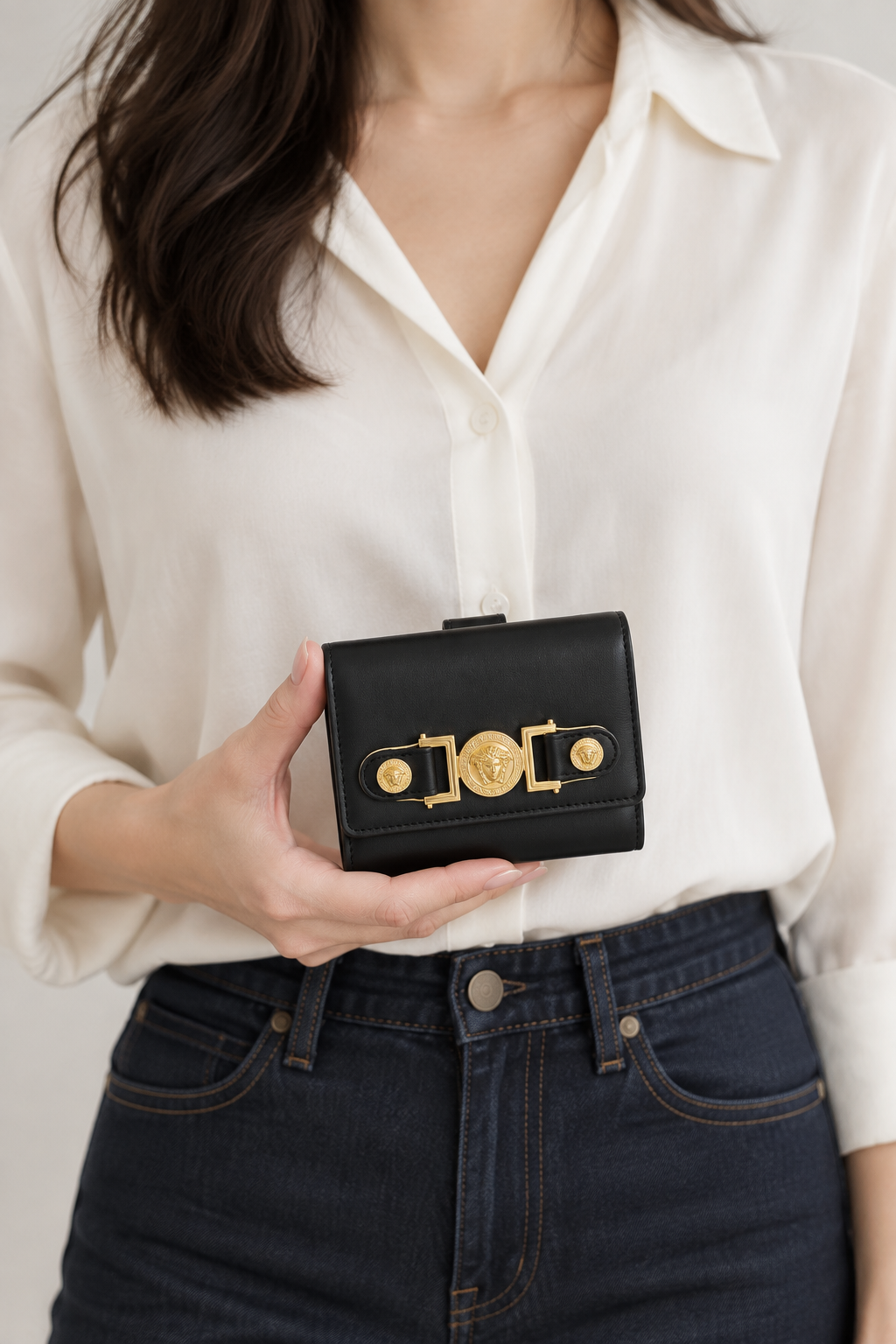 A woman holding versace medusa wallet wearing a white button-down shirt and dark blue jeans holds a small black leather clutch bag with a prominent gold-tone decorative clasp.
