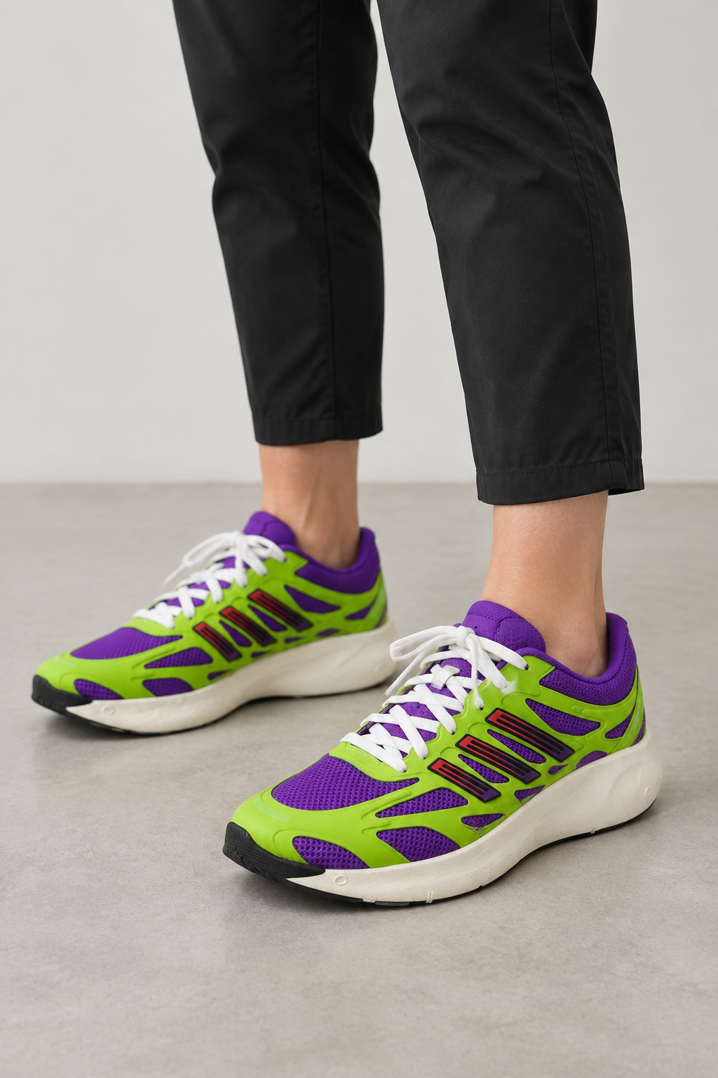 A person wearing bright neon green and purple running shoes with white laces and black pants, standing on a light gray floor against a light gray wall.
