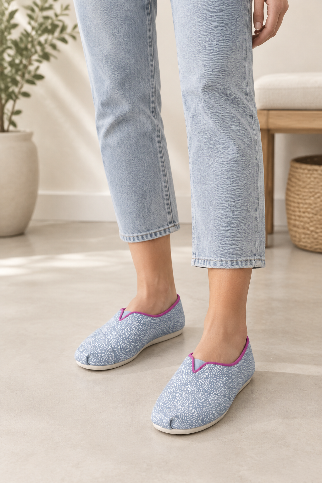A person wearing light blue cropped jeans and slip-on knit slippers with a blue-and-white speckled pattern and bright pink trim, standing in a bright indoor room.