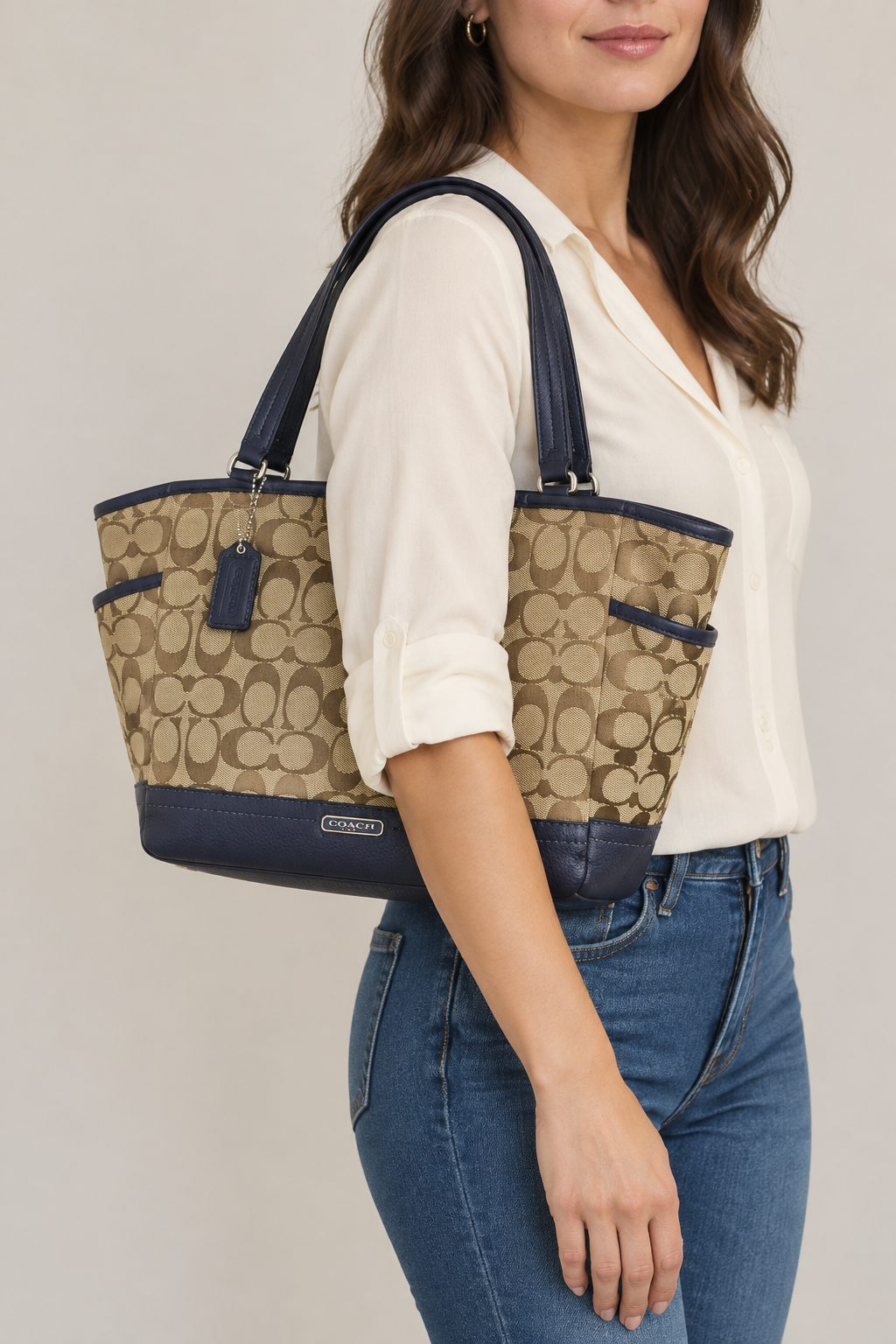 A woman wearing a white blouse and blue jeans carries a Coach tote bag with the brand's signature monogram pattern in beige and navy blue.