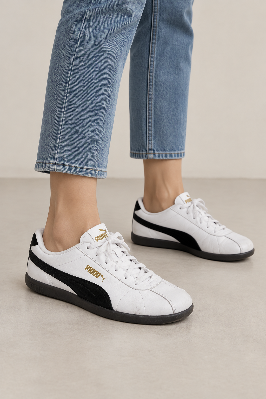 A pair of Puma sneakers in white, black, and gold, with one shoe standing upright and the other lying on its side to show the side profile.