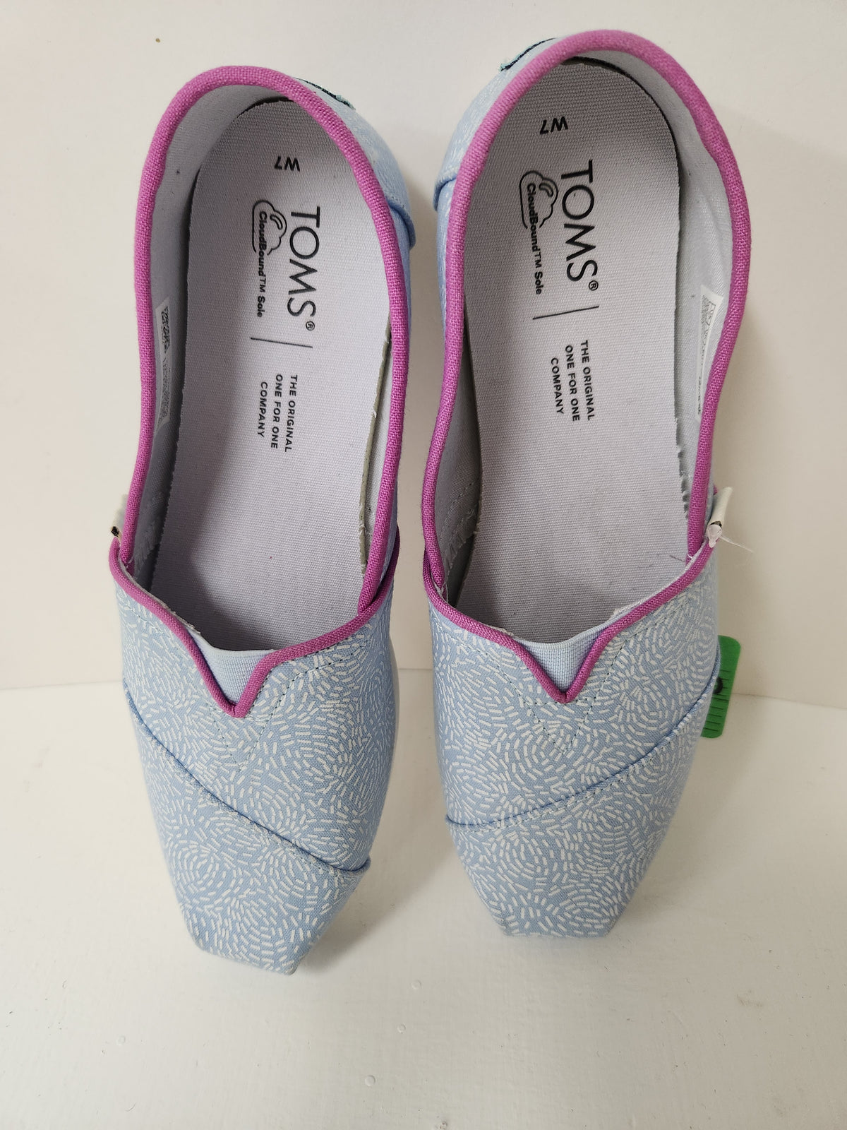 A pair of TOMS slip-on shoes in light blue with a white abstract pattern and bright pink trim, shown on a white surface.