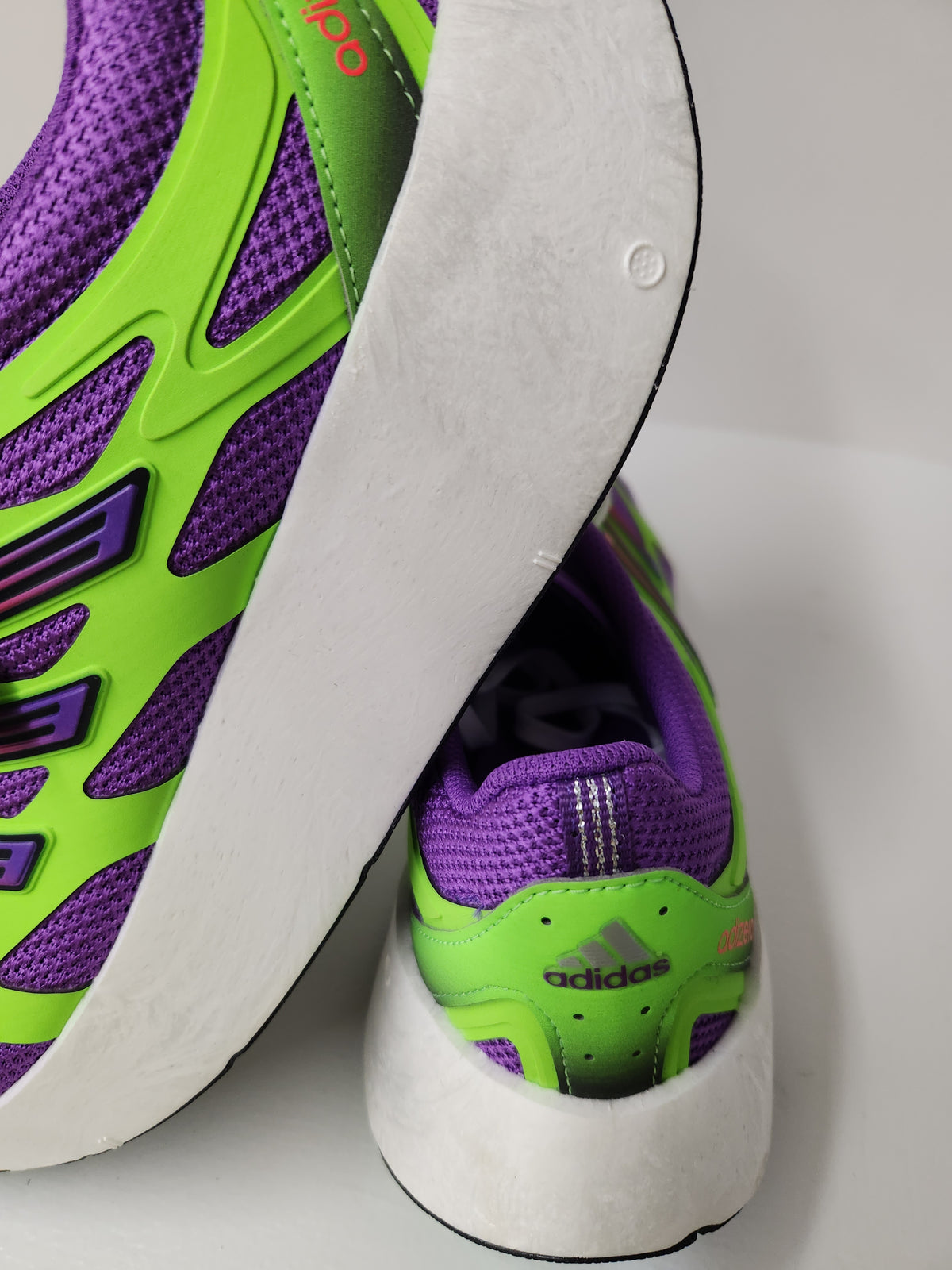 Close-up of an Adidas running shoe showing the side profile and heel, featuring a white midsole, neon green overlays, and purple mesh upper.