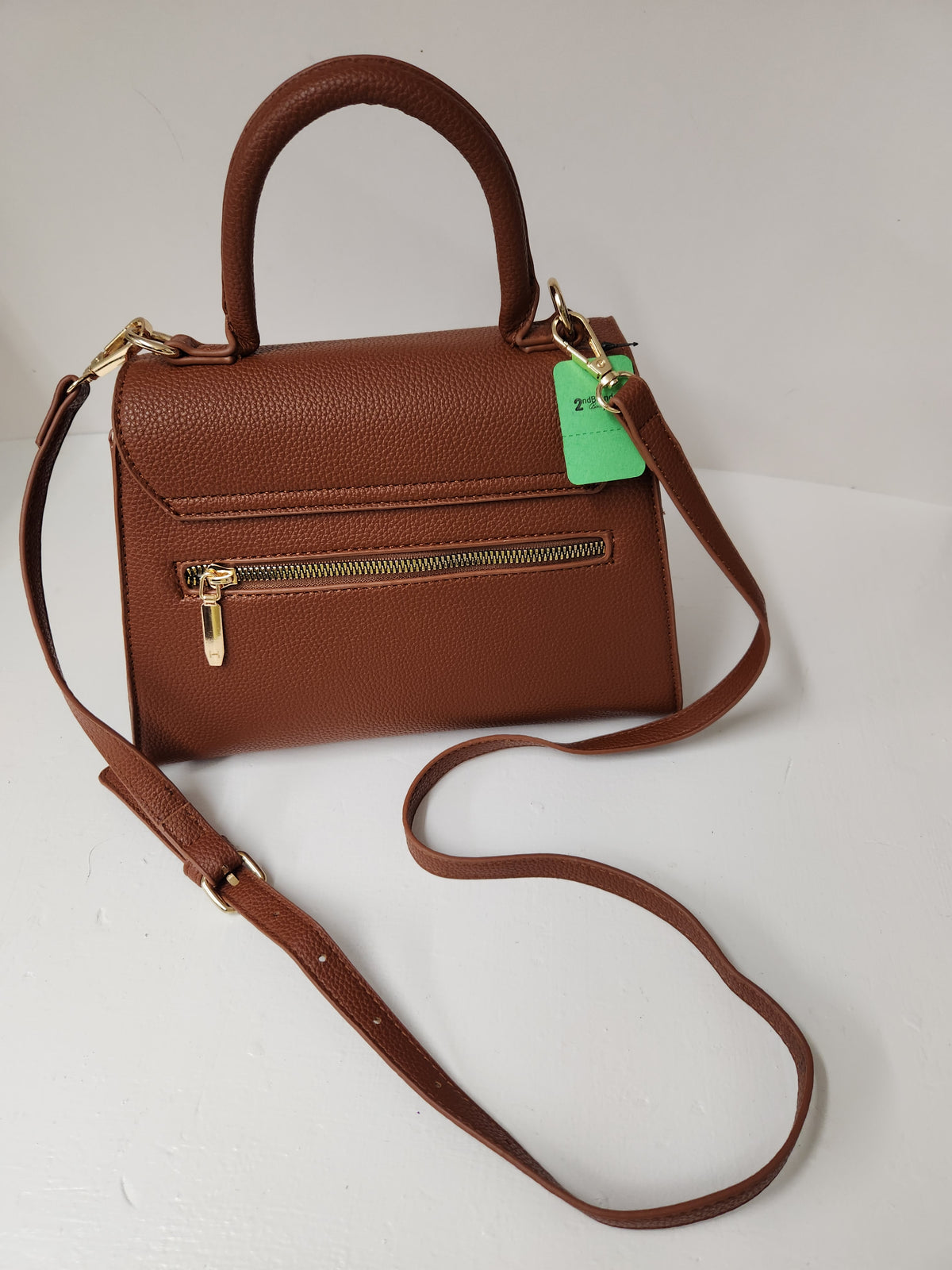 A brown pebbled leather handbag with a top handle, a front zip pocket, and a detachable shoulder strap, shown on a plain light background with a green hangtag attached.