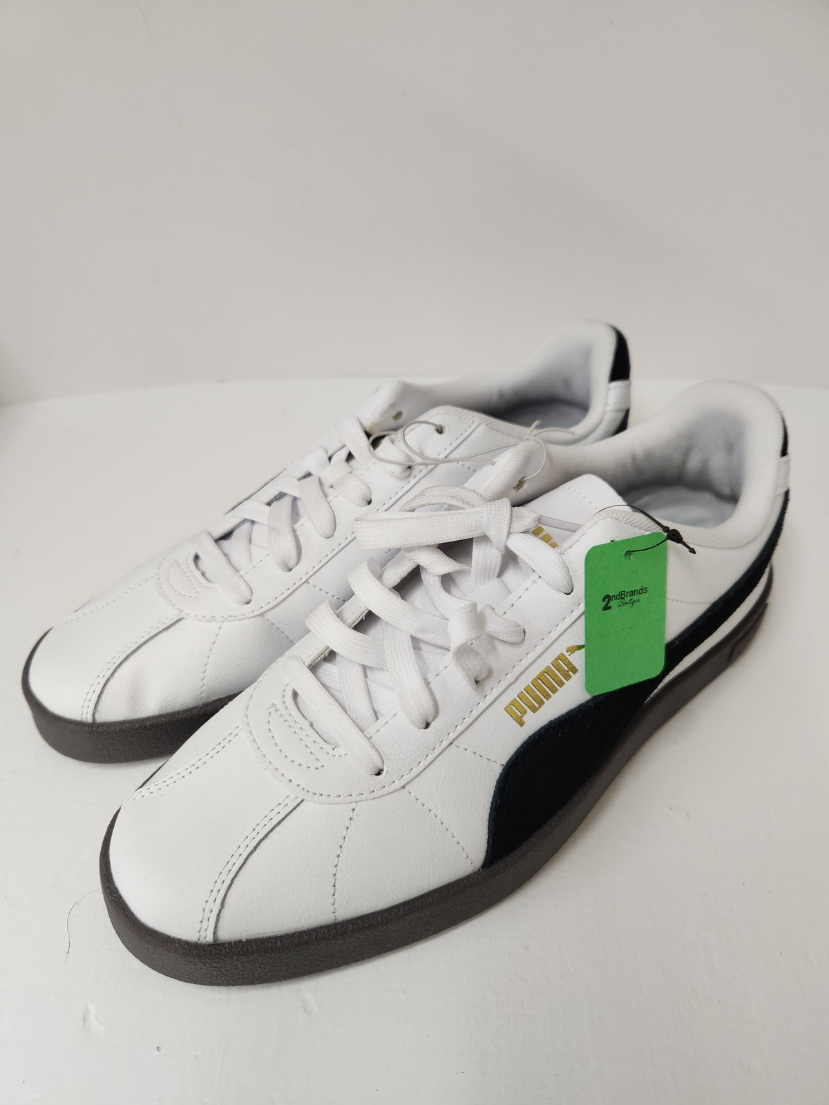 A pair of white Puma sneakers with black side stripes and a gold Puma logo, featuring a green resale tag on the side, set against a plain light background.