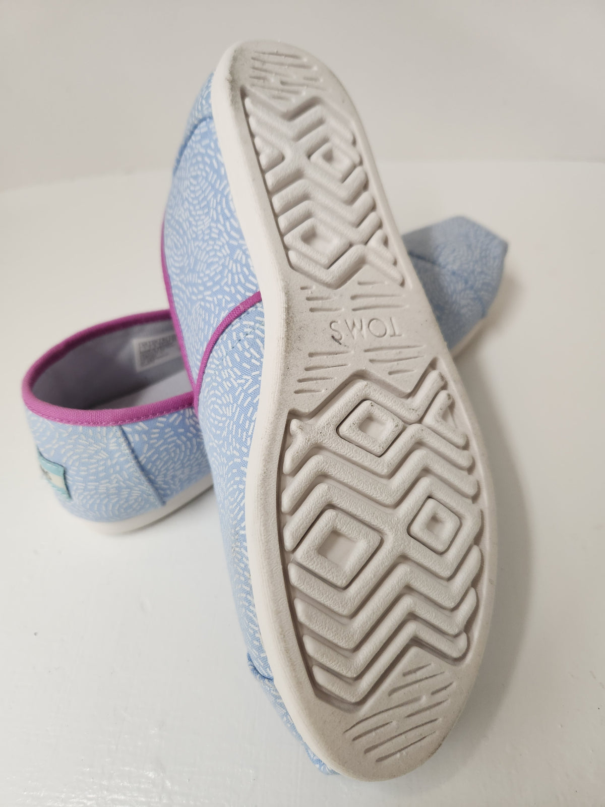 A pair of slip-on canvas shoes shown on a white surface, with one shoe standing upright to display the textured white outsole and the other lying down to show the light blue patterned upper and pink inner lining.
