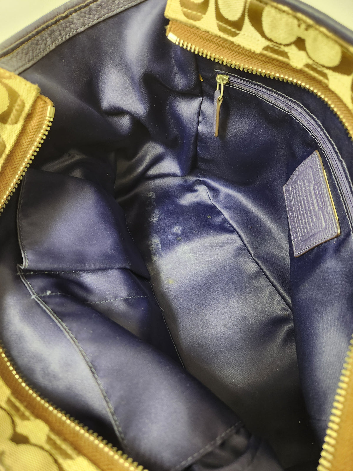 Close-up of the interior of a Coach handbag, showing a navy blue satin-lined compartment with gold-tone zippers and a gold patterned fabric lining.