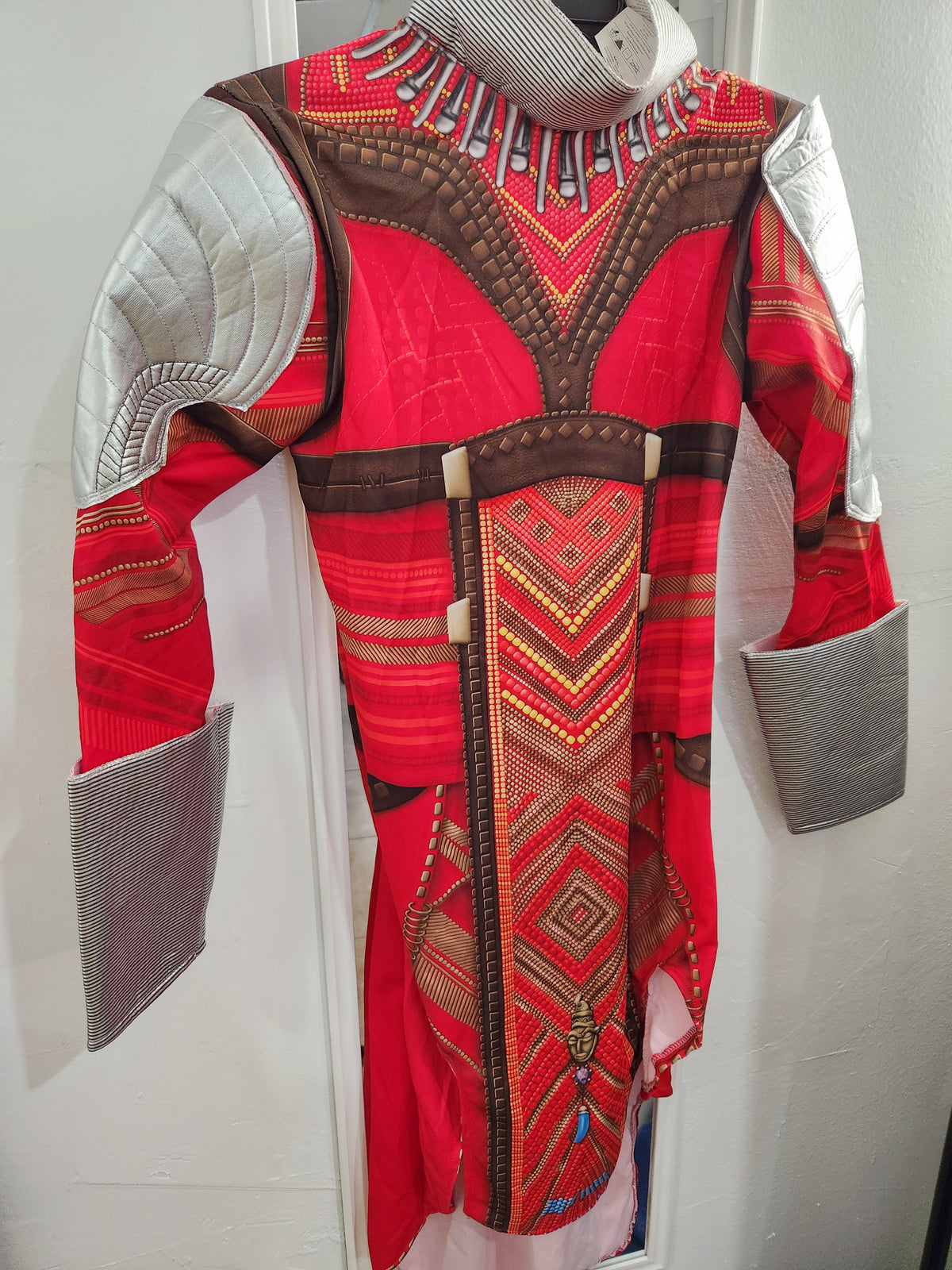 A red and brown patterned costume jacket or tunic with quilted silver-gray shoulder panels and matching cuff guards, displayed against a white wall. Wakanda's Dora Milaje Adult Costume