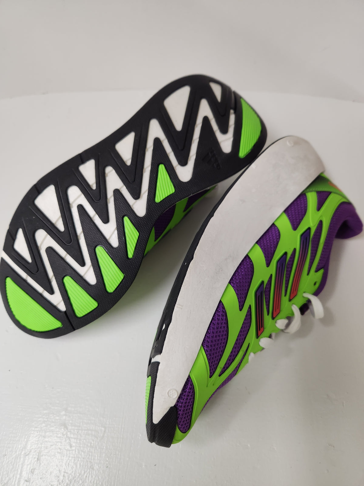 A pair of athletic sneakers shown from the side and sole views, featuring a bright green and purple mesh upper with a thick white midsole and a black outsole with a white zigzag pattern and neon green accents.