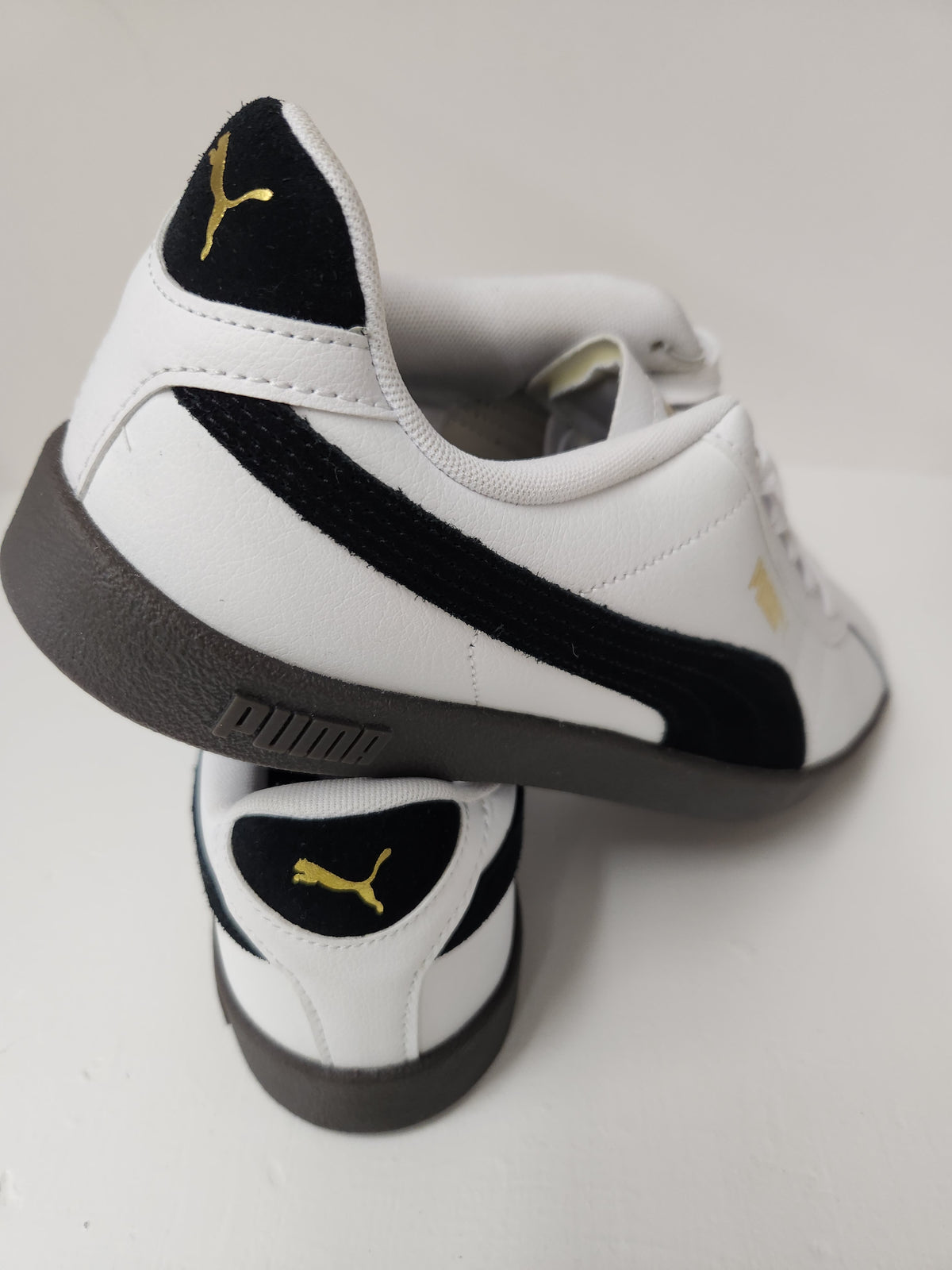 A pair of Puma sneakers in white, black, and gold, with one shoe standing upright and the other lying on its side to show the side profile.
