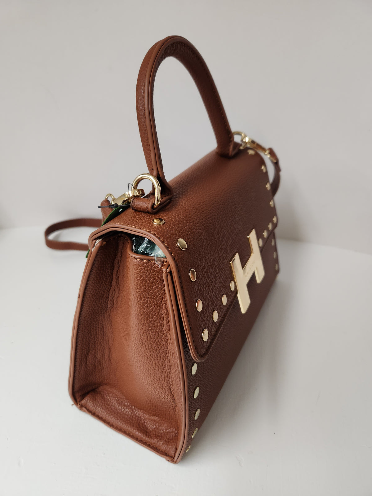 A brown pebbled leather handbag with a top handle, gold-tone studs, and a large gold 'H' logo on the front, photographed on a light gray background.
