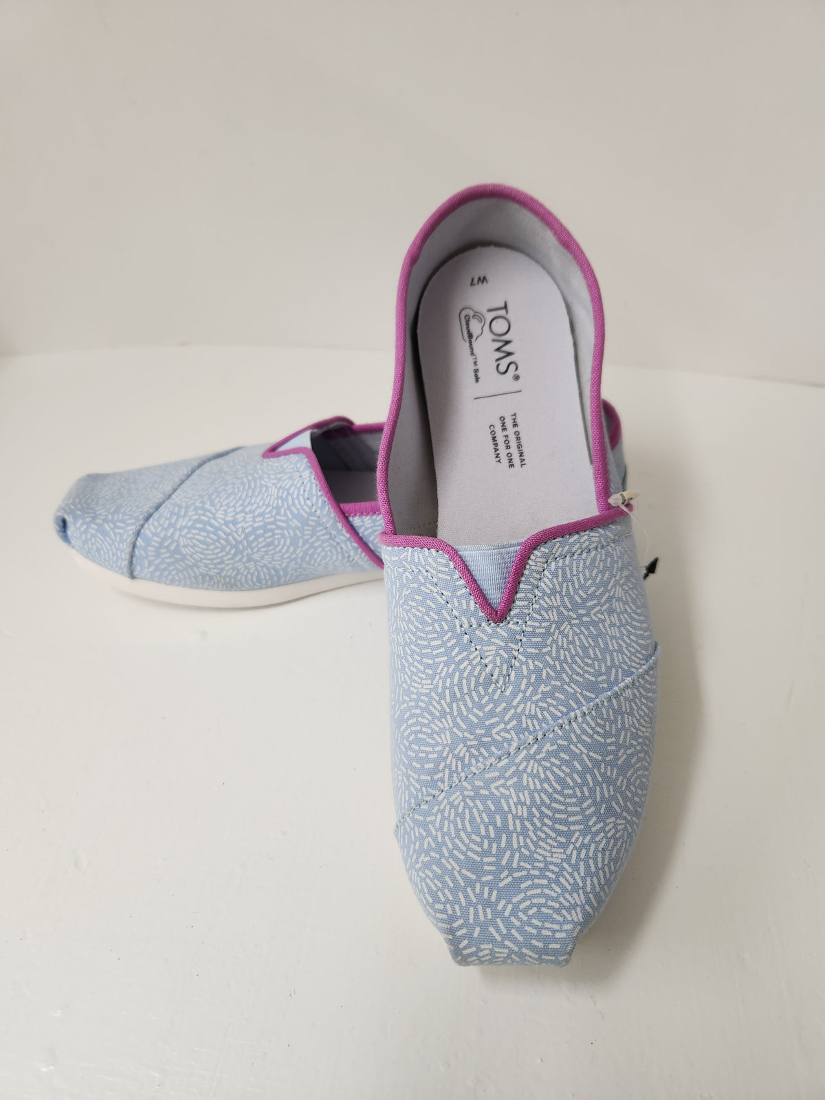 A pair of light blue Toms slip-on shoes with a white leaf pattern and magenta trim, displayed on a white surface with a white background.
