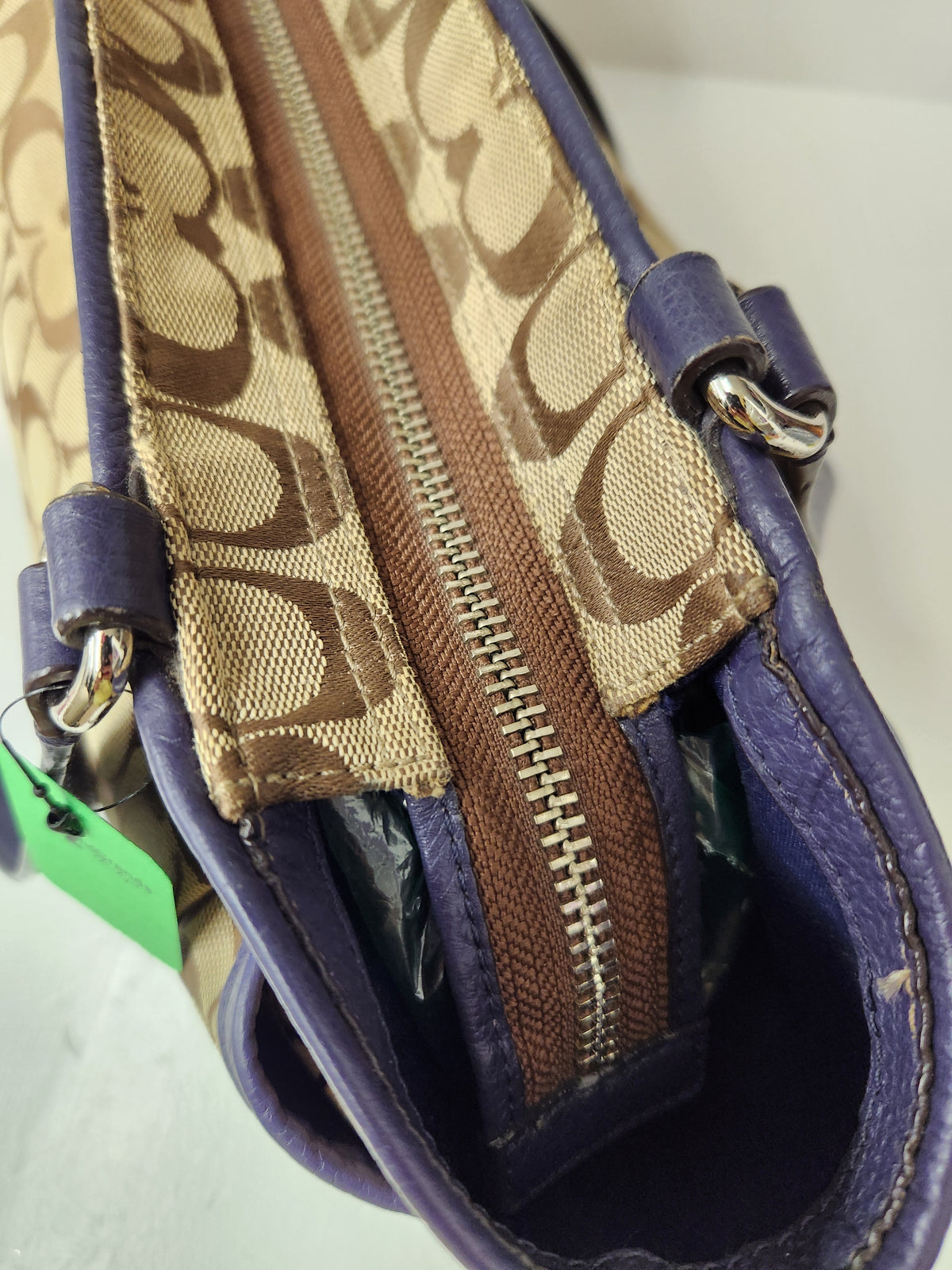Close-up of a Coach handbag showing the signature monogram canvas zipper track, purple leather trim, silver-tone hardware, and a green price tag.