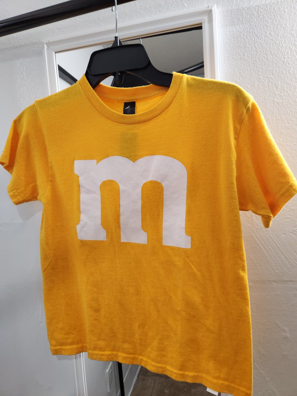 A yellow short-sleeve t-shirt hanging on a black hanger against a white door frame. The shirt features a large, bold white lowercase 'm' logo printed across the chest, representing the M&M's brand.