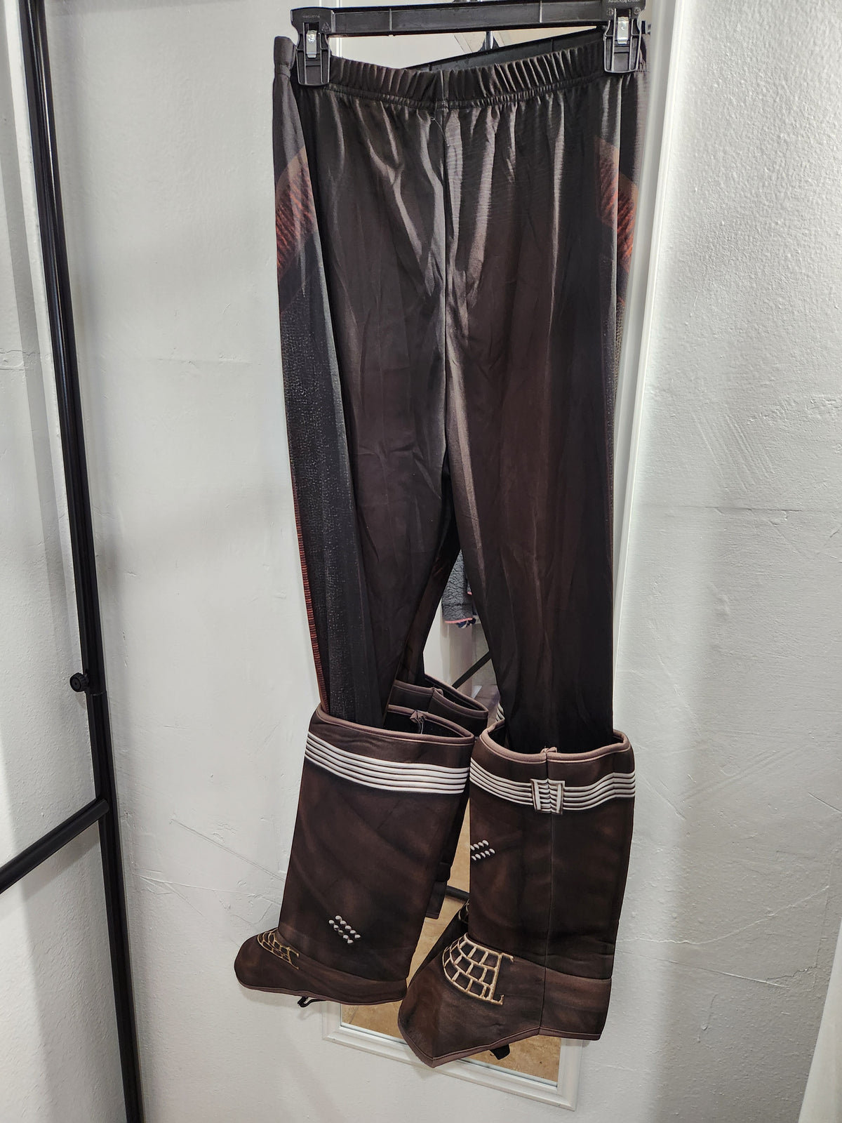 A pair of dark brown pants with matching tall brown boot covers hanging from a black hanger against a white wall.