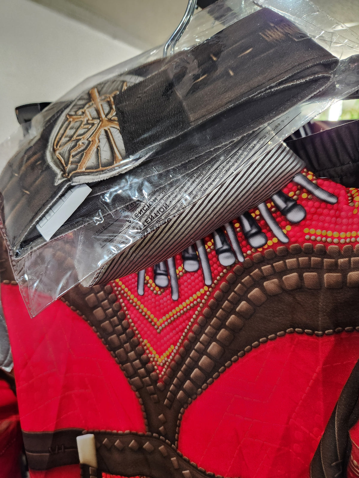 A close-up of a red and brown African dashiki garment with intricate beadwork and a matching black cap, both wrapped in clear plastic packaging.