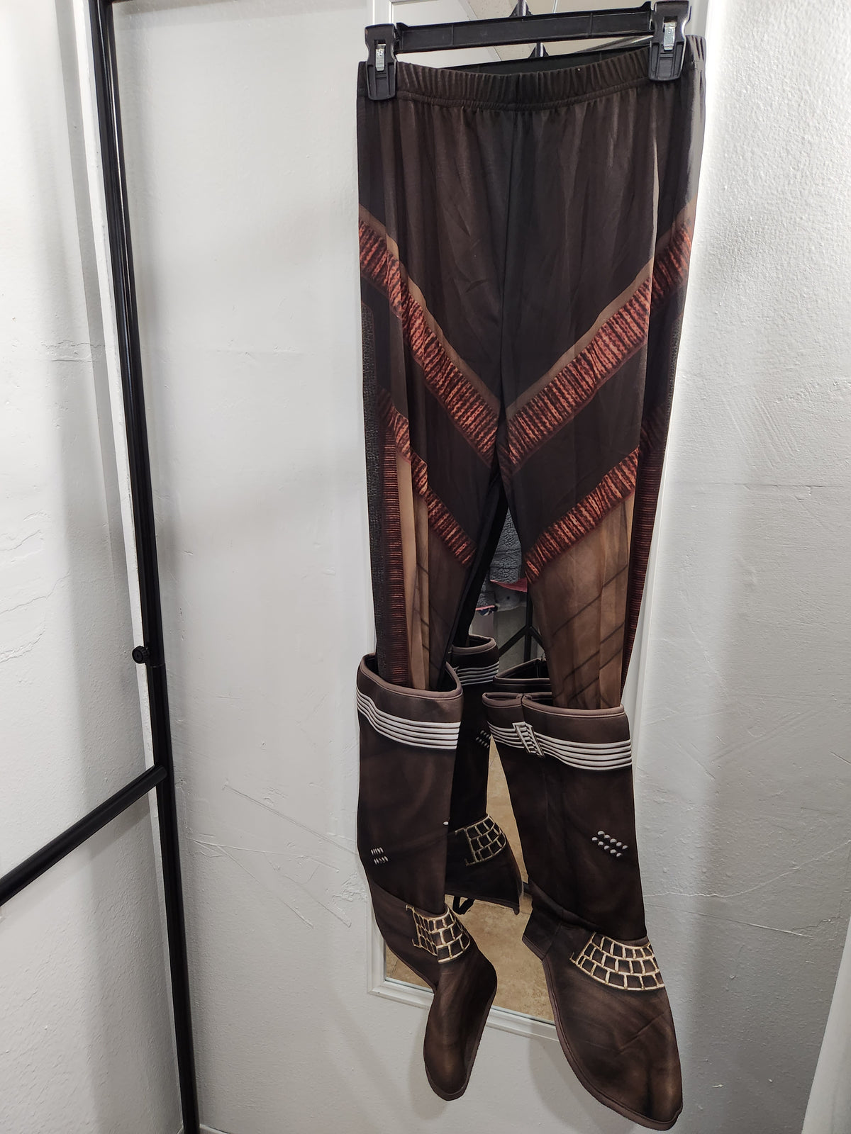 A pair of dark brown sheer leggings with diagonal red and orange geometric stripes, paired with matching dark brown knee-high boots featuring white stripes and textured details, hanging on a black hanger against a white wall.