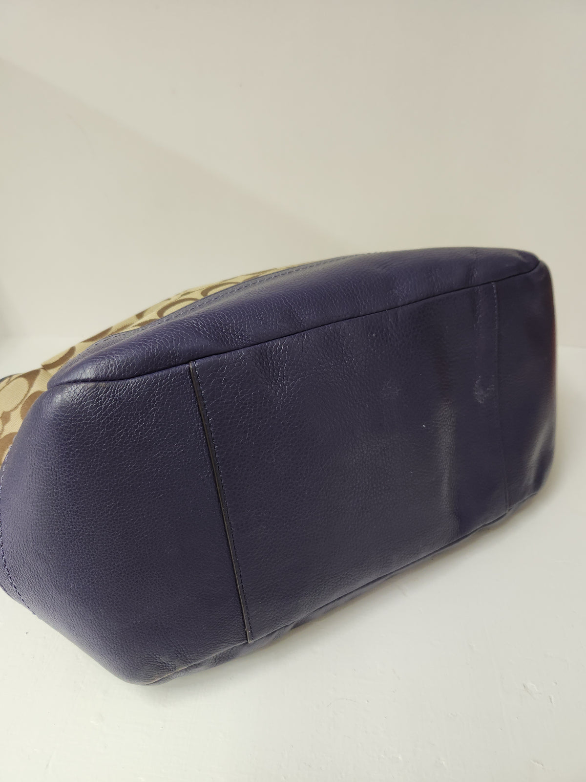 A Coach handbag in navy blue pebbled leather with the brand's signature beige and navy monogram canvas partially visible on the side, photographed on a plain off-white background.