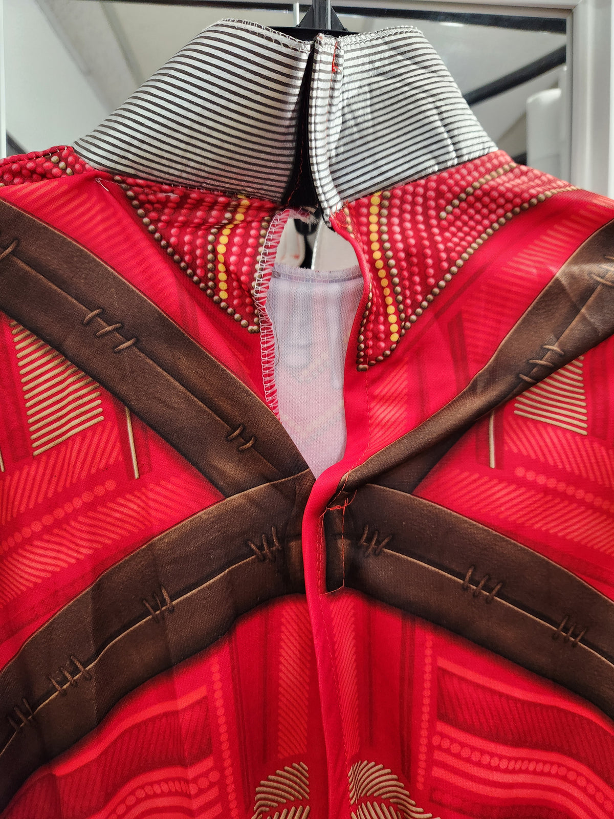 A close-up of a red patterned garment with brown cross-shaped panels, a striped collar, and decorative stitching details, shown hanging on a hanger.
