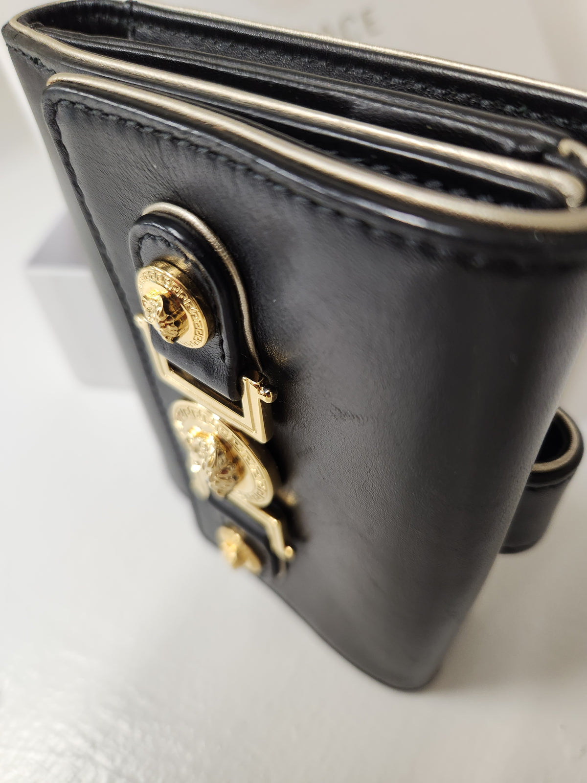 A close-up of a versace black leather wallet featuring ornate gold-tone hardware and decorative studs on a light gray surface.